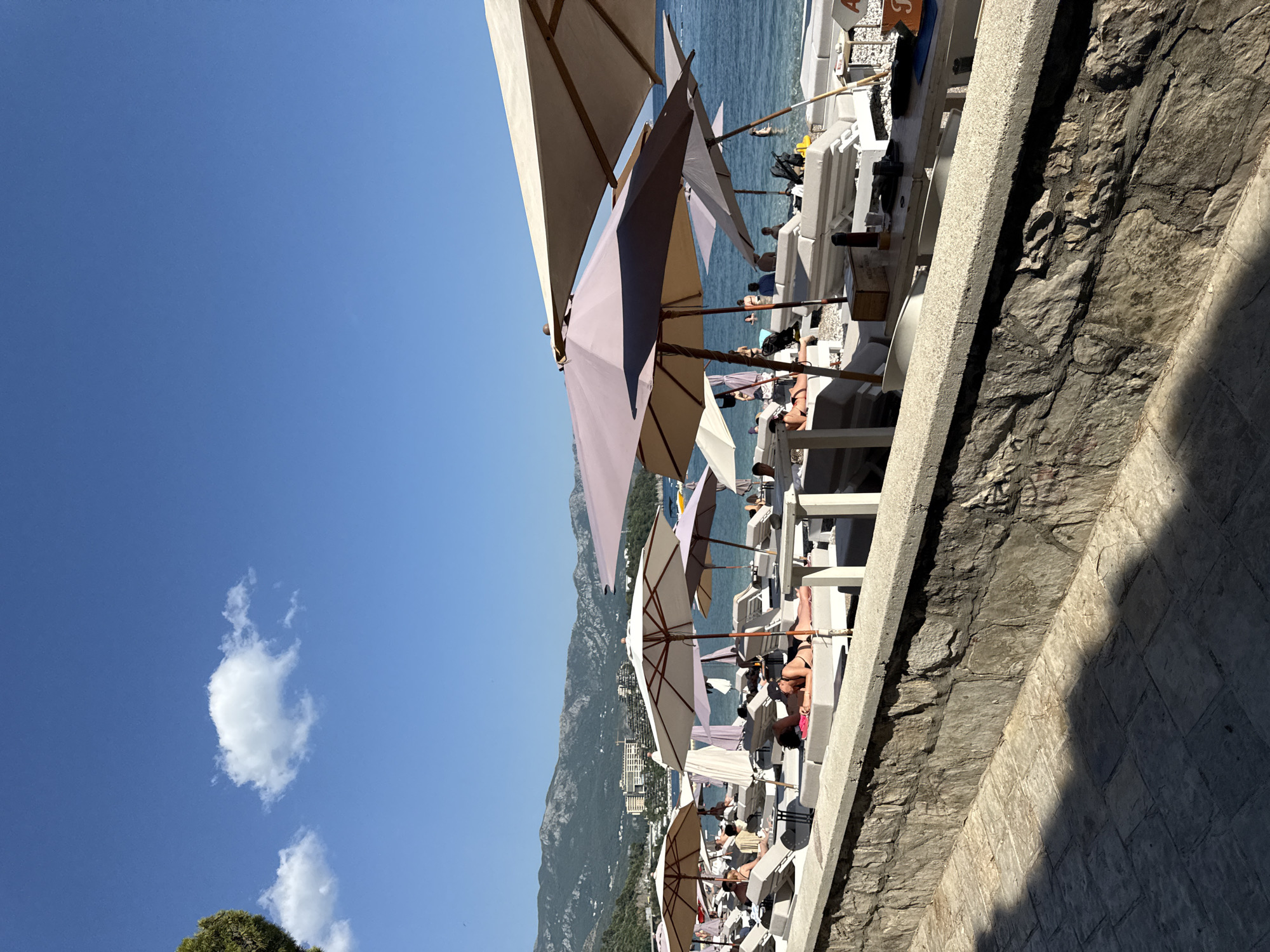 The image depicts a sunny day at a beachside resort, likely in Budva, Montenegro. The scene is dominated by rows of white lounge chairs and umbrellas set up on a pebbled beach, with the blue sea and a lush green mountainous backdrop. Several people are visible, relaxing on the lounge chairs, some wearing swimwear. The atmosphere appears tranquil and leisurely, suggesting a vacation or a day of relaxation.
Umbrellas of varying colors, including beige, purple, and white, provide shade for the beachgoers. The lounge chairs are neatly arranged, implying a well-maintained establishment. In the background, the clear blue sky with a few scattered white clouds indicates fair weather. The sea is calm and inviting, with a few people visible swimming or enjoying the water. The mountainous terrain behind the beach suggests a coastal Mediterranean setting.
The time of day appears to be daytime, given the bright sunlight casting shadows, and the overall luminosity of the scene. The presence of lounge chairs, umbrellas, and people in beach attire clearly points to a beach resort or a similar recreational area. There are no immediately obvious signs of specific events or activities beyond general relaxation and enjoyment of the beach and sea. The overall mood is one of peacefulness and summer enjoyment.