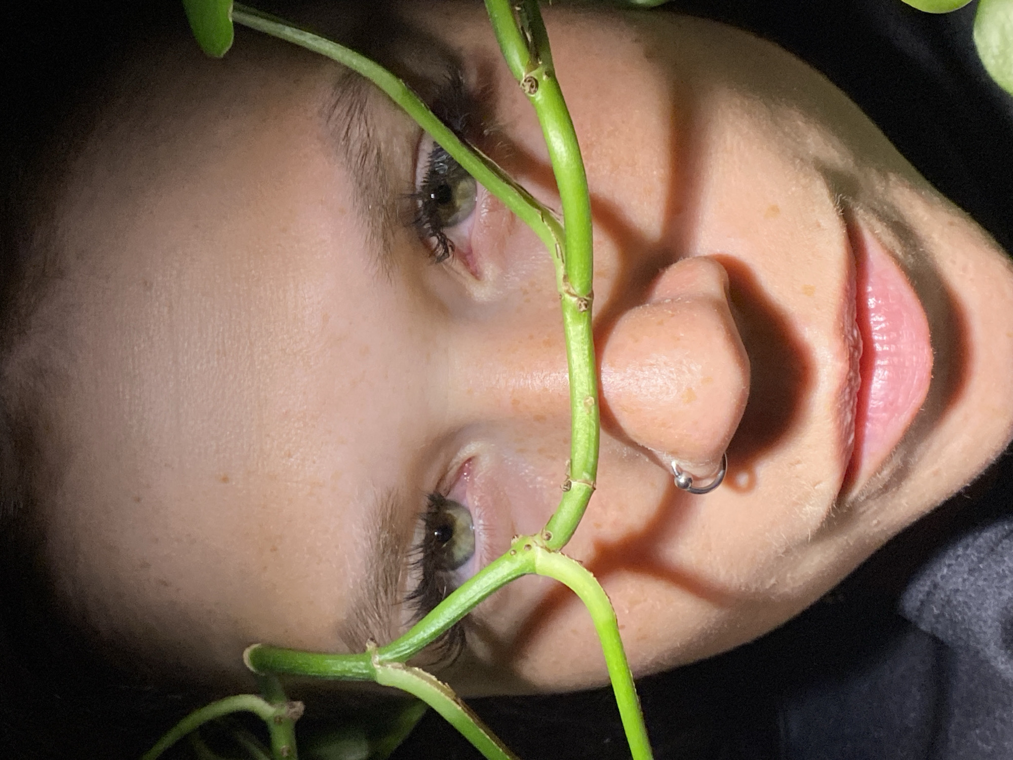 A close-up, low-angle shot focuses on the face of a person, framed by a green vine. The individual has light skin with visible freckles, green eyes, and dark eyelashes. A silver nose ring is visible on their right nostril. The vine is draped across the face, obscuring parts of the eyes and nose. The background is dark, suggesting artificial lighting. The setting is not identifiable as being in the city of Chaikovsky, Russia.