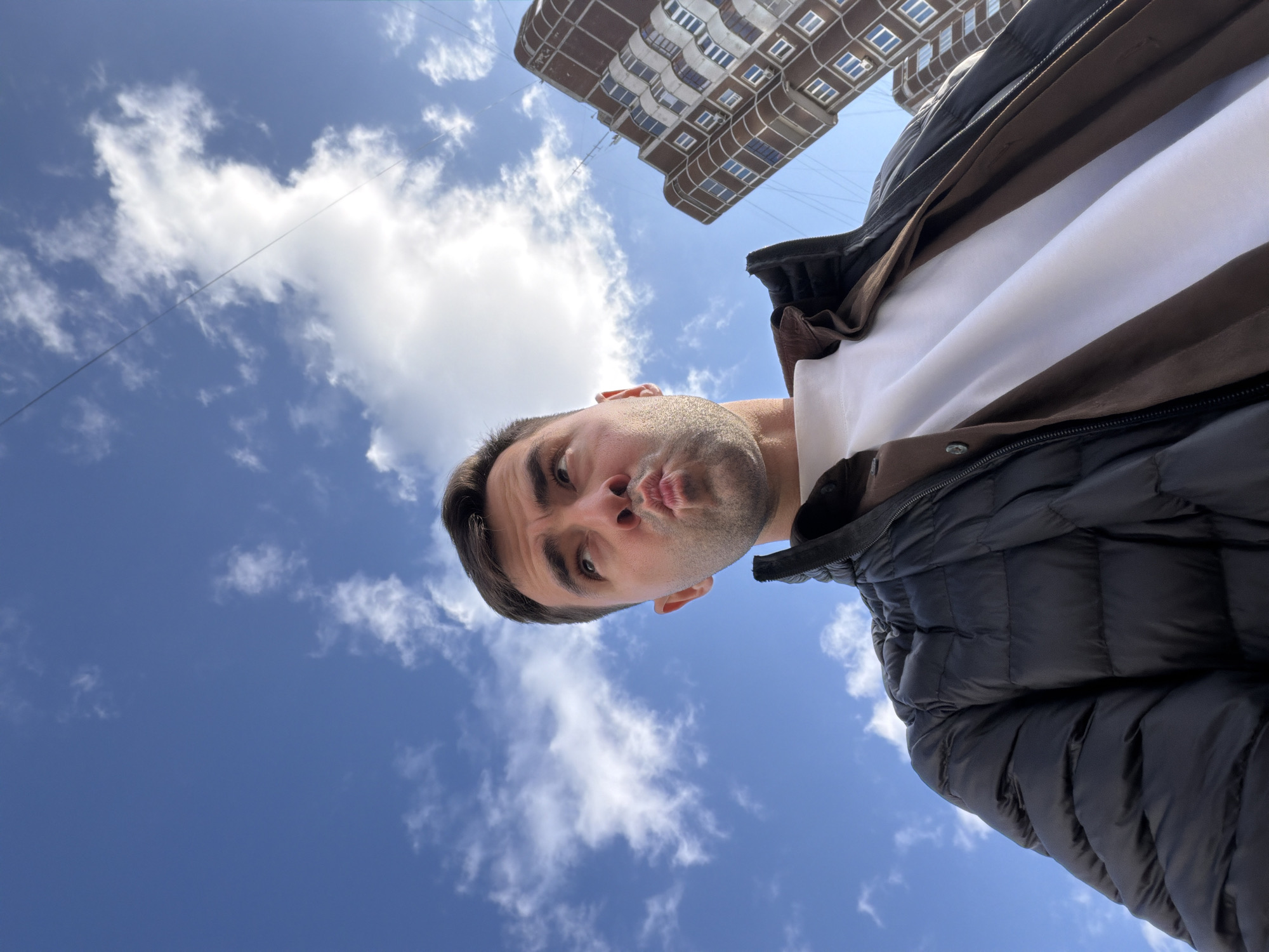 A low-angle shot shows a man with short dark hair, light skin, and a pouting expression. He wears a white t-shirt under a brown and white jacket and a black puffer jacket. Behind him is a blue sky with scattered white clouds. A multi-story building with rows of windows stands in the background. The scene suggests an outdoor location in an urban setting. There is no specific information in the image to indicate the exact location in Krasnoyarsk, Russia.