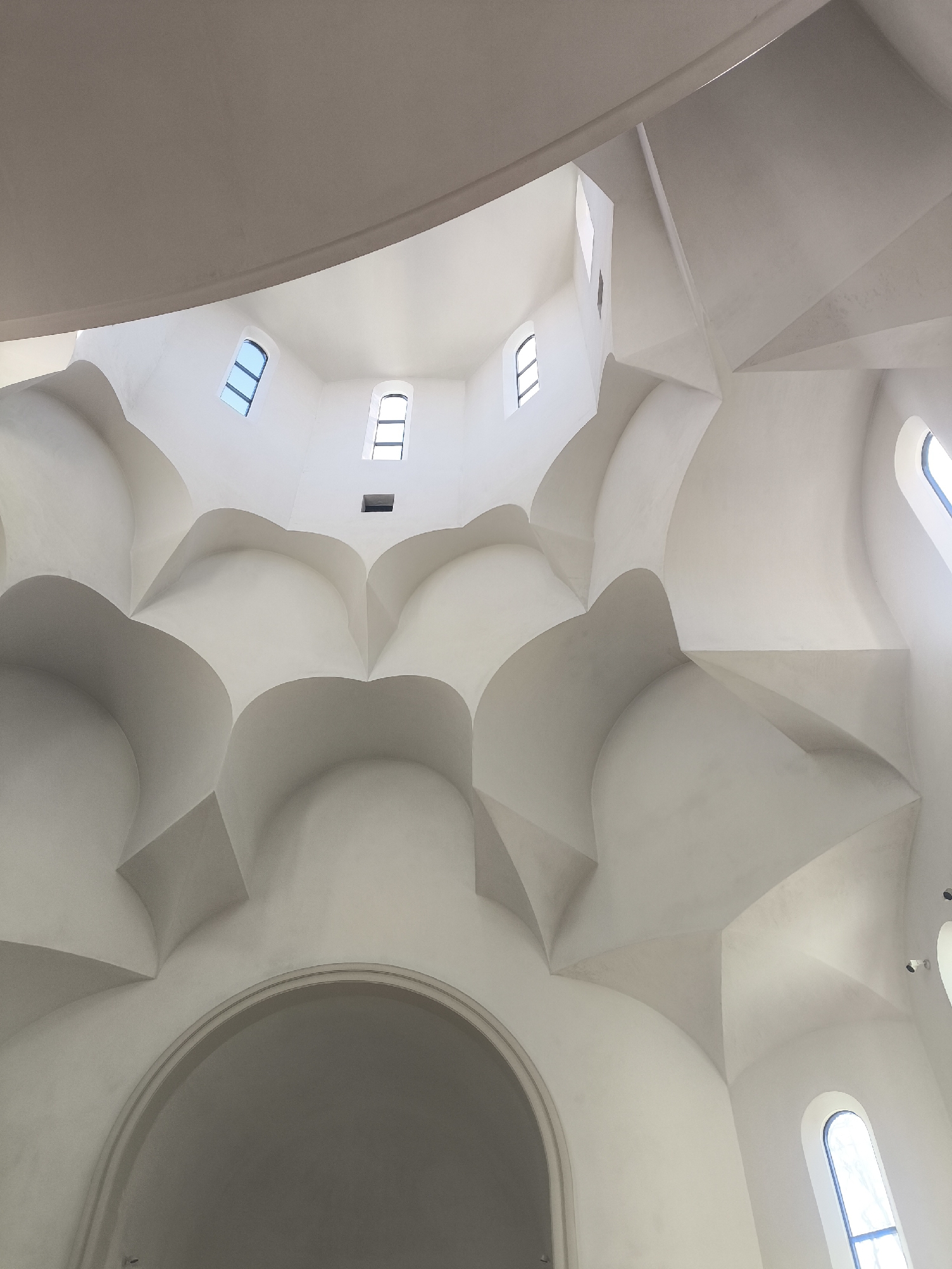 The image showcases the interior ceiling of the Saint Paul Catholic Church located in Strasbourg, France. The ceiling's architecture is a complex arrangement of interconnected, rounded, white plaster sections that resemble an intricate, symmetrical pattern of domes or arches. Towards the top, several rectangular windows are visible, letting in natural light. An arch is visible near the bottom of the frame. Some lighting fixtures are visible on the right side.