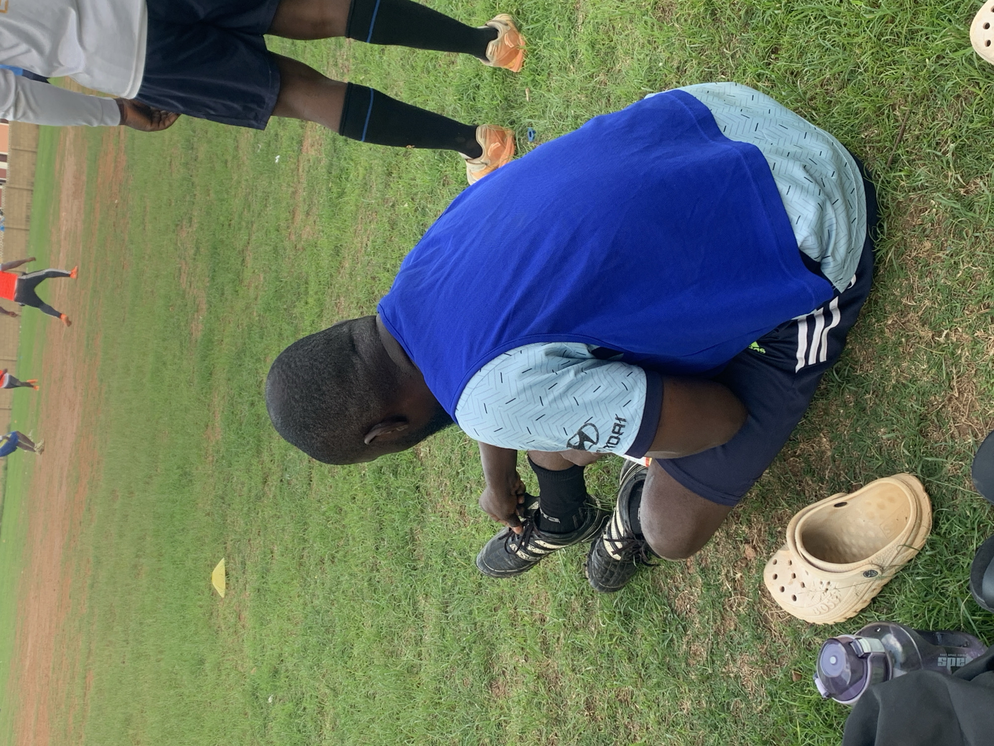 A man is kneeling on a grassy field, tying the laces of his black athletic shoes. He is wearing black socks, navy blue shorts with white stripes, a light blue short-sleeved shirt with a subtle patterned design and the word "HYUNDAI" printed on the sleeve, and a bright blue sleeveless top. Next to him on the grass are a tan-colored clog, a water bottle, and a black bag. In the background, other people are on the field. The scene appears to be in Jos, Nigeria.