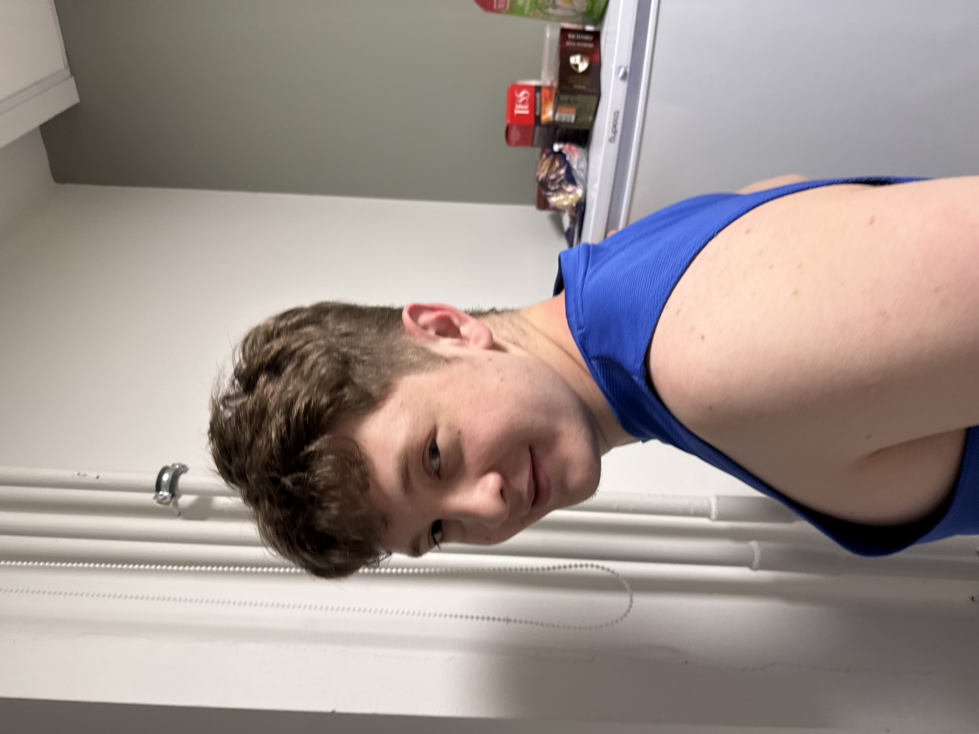 A young man with short brown hair and wearing a blue tank top is shown in the image. The setting appears to be indoors, possibly a kitchen or pantry area, with a white wall and a set of pipes running horizontally along the wall. In the background, there is a refrigerator with various food items and boxes placed on top. The image is rotated, giving the impression that the man is lying down or leaning backwards. There is no information to confirm the location of the photo as being in Kazan, Russia.
