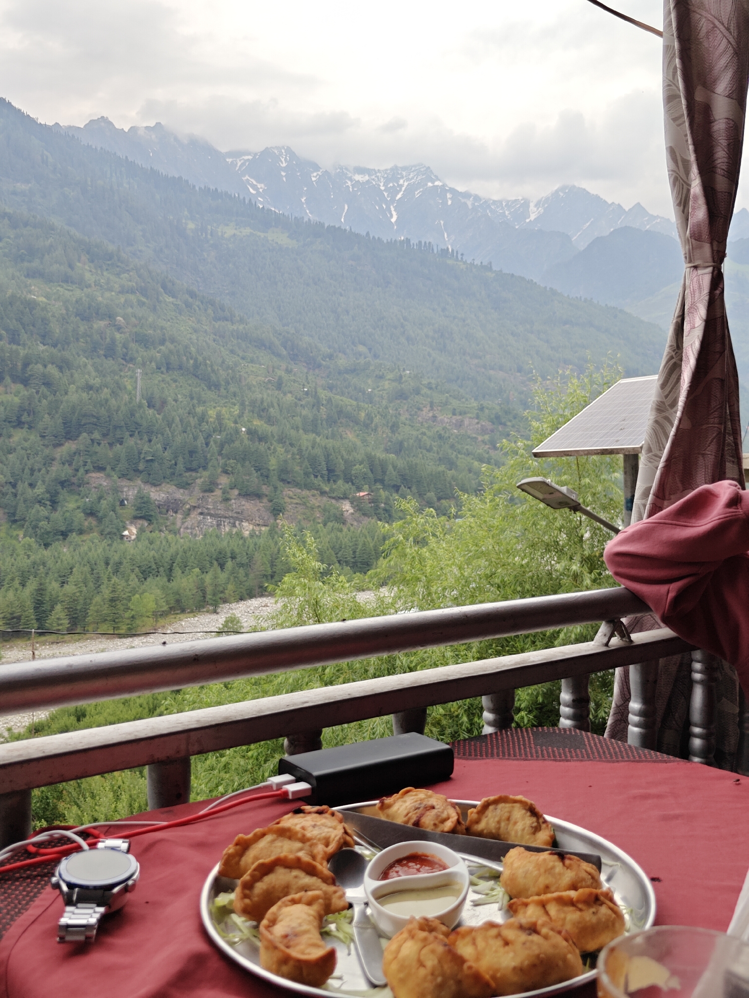 The image captures a scenic view from a balcony in Vashisht, India, overlooking a lush green valley with snow-capped mountains in the distance. On the table, a plate of fried dumplings, a small bowl with two dipping sauces, and a watch are visible. A power bank and charging cables are also present. A person wearing a pink hoodie is partially visible on the right side of the frame, leaning on the railing. The atmosphere appears to be overcast, with the mountains shrouded in mist.