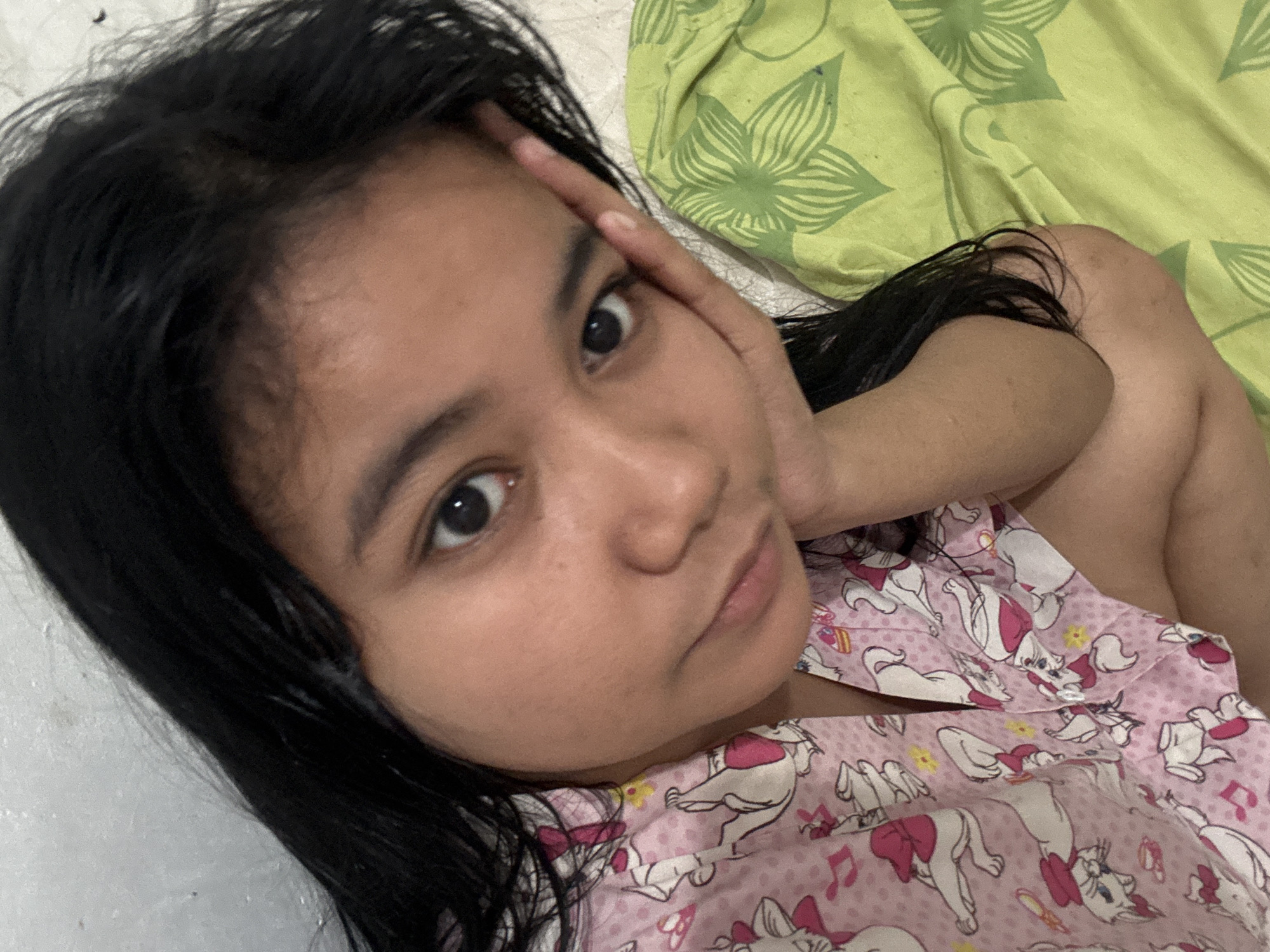 A young woman with long, dark hair is resting her cheek on her hand. She is wearing a pink pajama top with a repeating pattern of white cats with pink bows. The background consists of a white wall and a green cloth with a floral pattern. The scene appears to be indoors, possibly in a bedroom, in North Jakarta, Indonesia.