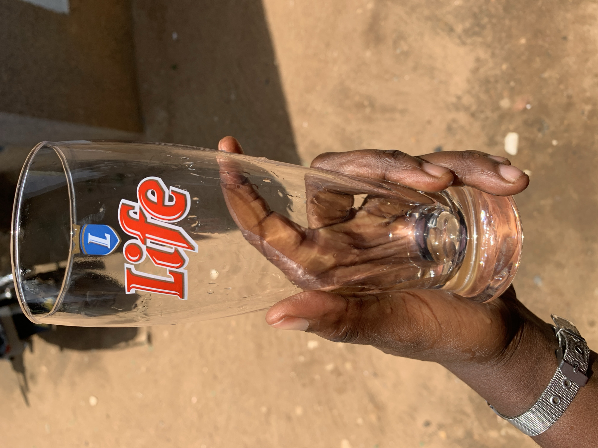 The image, originally rotated 90 degrees, features a dark-skinned hand holding a clear glass horizontally against an outdoor, sunlit background. The hand, an adult's, firmly grips the glass. Water droplets or condensation are visible on the surface of the glass, suggesting it recently contained a cold drink.
On the glass, there is a prominent red and white logo spelling "Life," accompanied by a blue shield emblem above it that appears to read "1 L" (likely indicating a one-liter capacity). A silver-colored mesh watch with a buckle is worn on the wrist of the hand holding the glass.
The background shows a light brown, textured ground surface, possibly dirt or sand, consistent with an outdoor environment. Strong, high-angle sunlight is evident, casting sharp shadows and brightly illuminating the subjects, indicative of a clear, sunny day, likely around midday. The scene is set in the context of the New Stadium, Nigeria. The image captures a simple moment of a person holding a glass, possibly enjoying a beverage, in an outdoor setting.