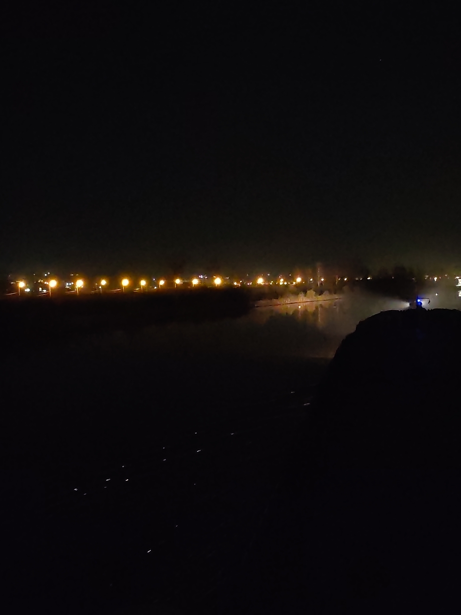 The image depicts a night scene in Yakhroma, Russia. A row of streetlights illuminates a distant waterfront, their reflections visible in the water. A dark, silhouetted structure is visible in the foreground, likely a rocky outcrop or a building, while a blue light, presumably from a vehicle, shines from the right side of the image. A few faint streaks of light are present near the bottom.