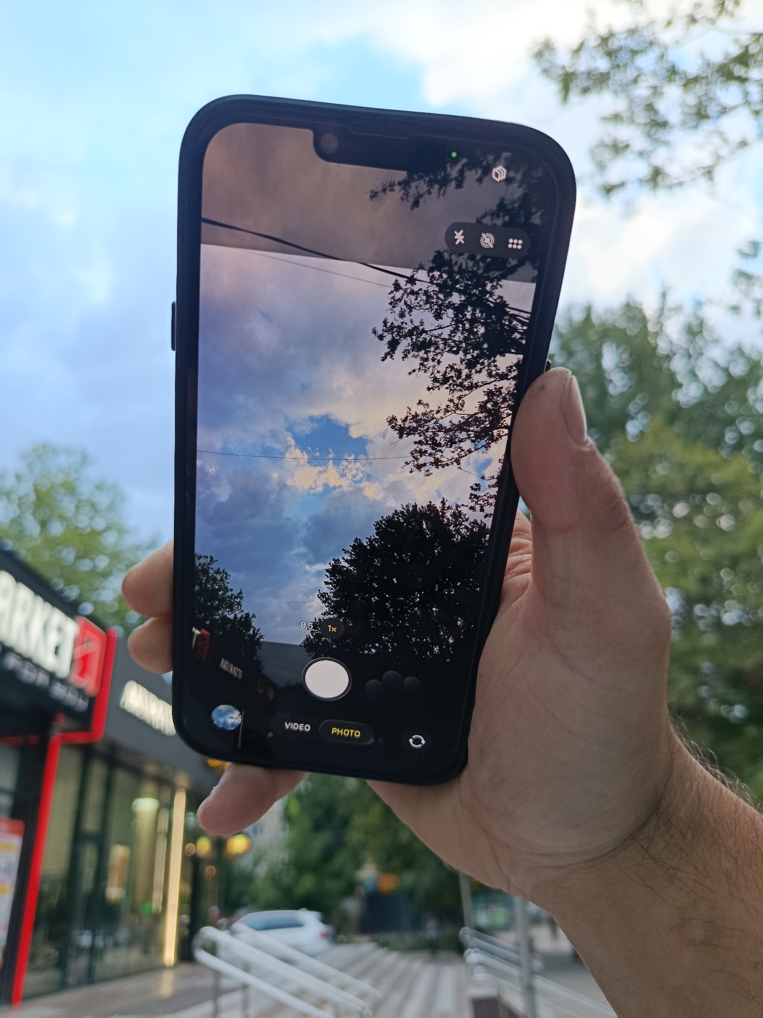 A hand holds a smartphone displaying a camera interface, framing a cloudy sky and silhouetted trees. The phone screen shows camera controls, including options for video and photo, and zoom levels. In the background, a blurred urban scene features a building with signage that appears to read "MARKET FOR MEN," suggesting a commercial area in Chișinău, Moldova. The overall lighting indicates it is daytime, likely overcast or late afternoon.