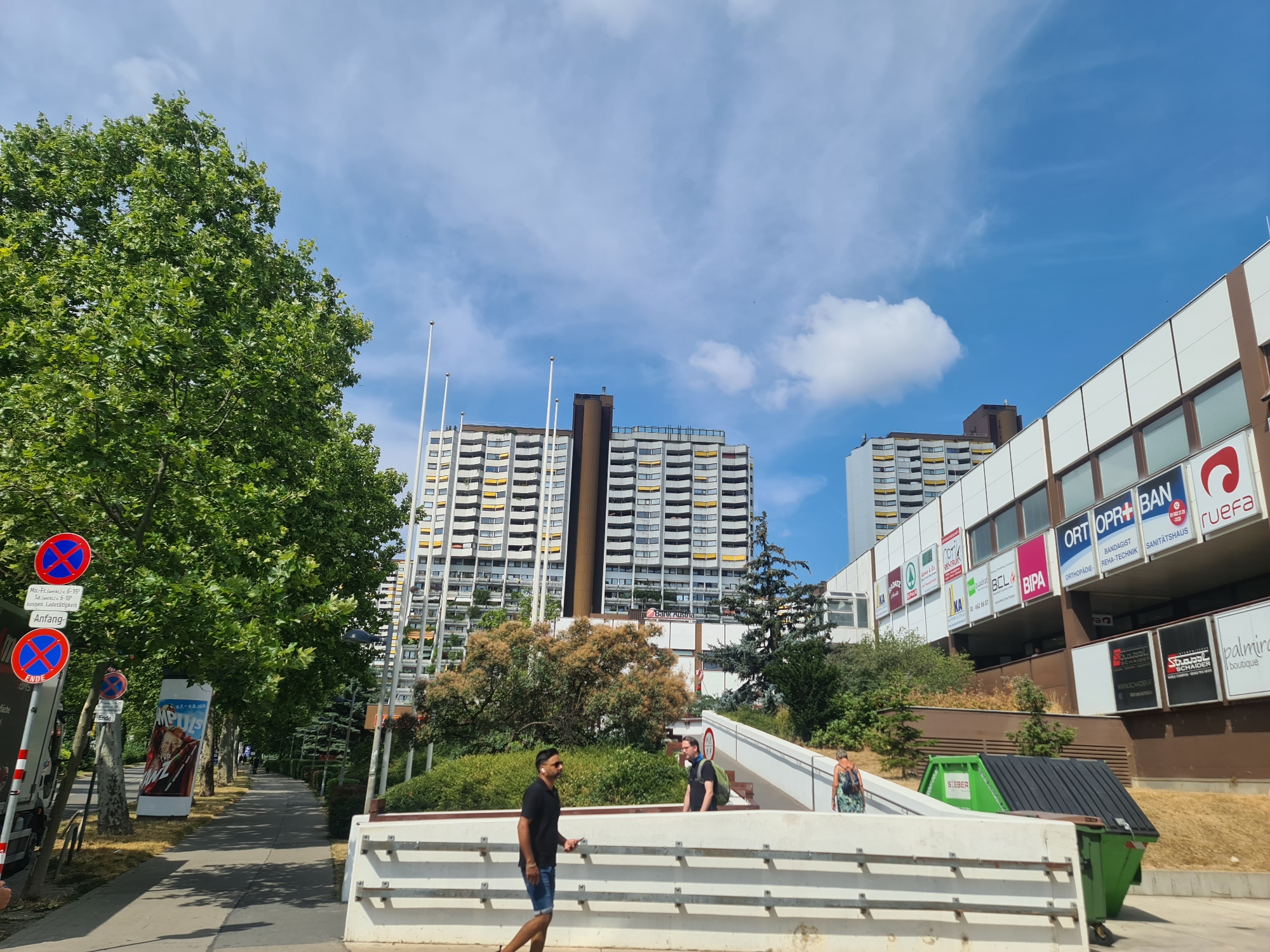 This is a wide shot of a pedestrian walkway and buildings in Vienna, Austria, on a sunny day with a blue sky and scattered clouds. The walkway is lined with trees on the left, and a large, multi-story apartment building with white balconies and yellow accents dominates the background. To the right of the apartment building, a commercial building with various signs and a slanted facade is visible. Several people are present: one man in a black shirt and shorts walks away from the camera on the walkway; another man with a backpack stands on a white staircase; and a woman with a backpack walks up the staircase. Two green and brown trash bins are situated near the commercial building. Signs with red circles and diagonal lines, indicating parking restrictions, are posted on the left side of the walkway, along with a movie poster.