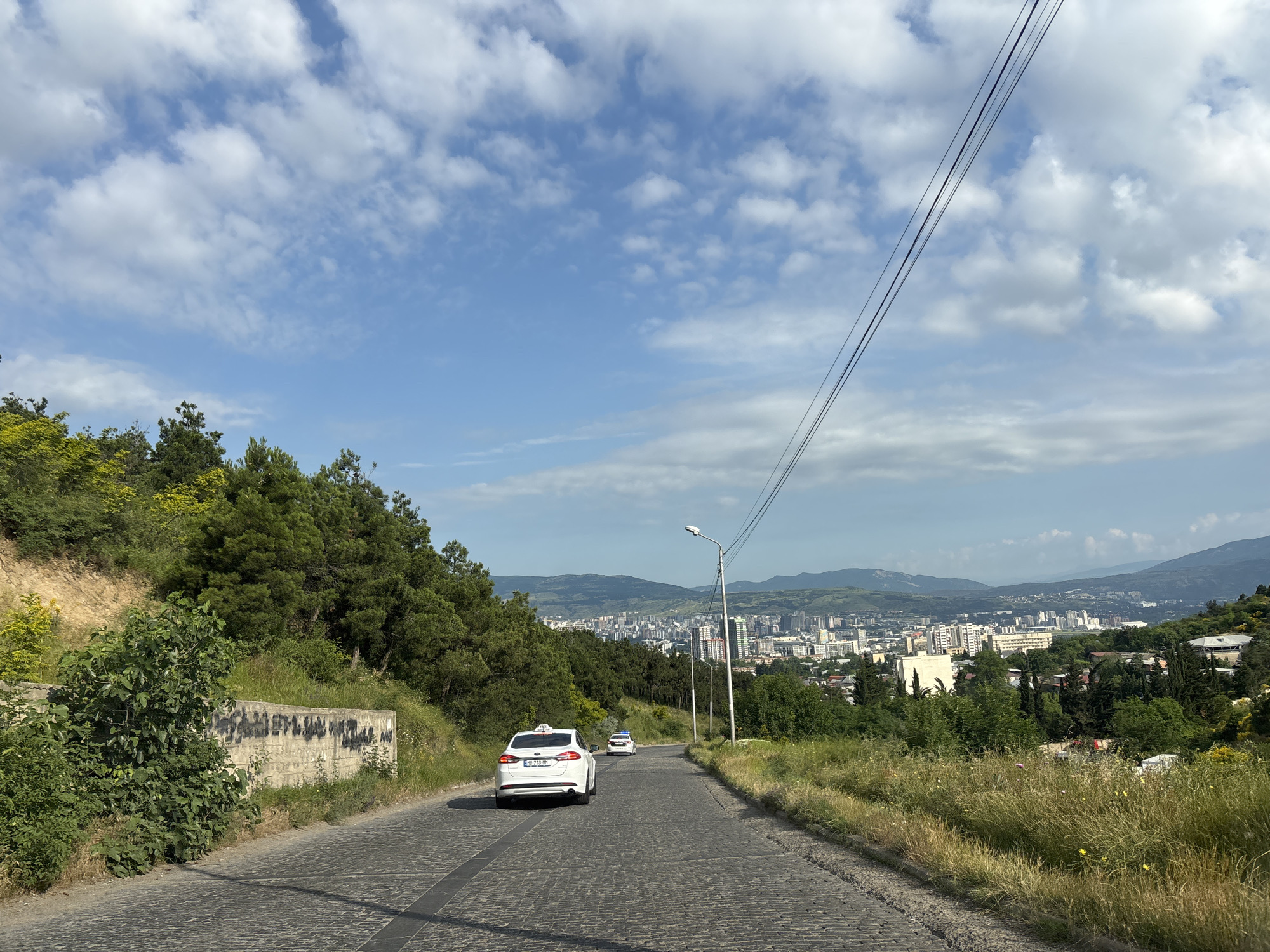 The image shows a view from a street overlooking the city of Tbilisi, Georgia. The street is paved with cobblestones and curves gently downhill. On the left side of the street, there is a steep, tree-covered hillside with a concrete wall at its base. On the right side, there is a grassy slope with wildflowers. In the distance, the city of Tbilisi spreads out across a valley, with numerous buildings of various heights and architectural styles. Mountains can be seen in the background. Two white cars, possibly taxis, are visible on the street, one in the foreground and another further up the road. Overhead, power lines stretch across the frame, and a street lamp is visible on the right side of the road. The sky is partly cloudy with fluffy white clouds against a blue background.