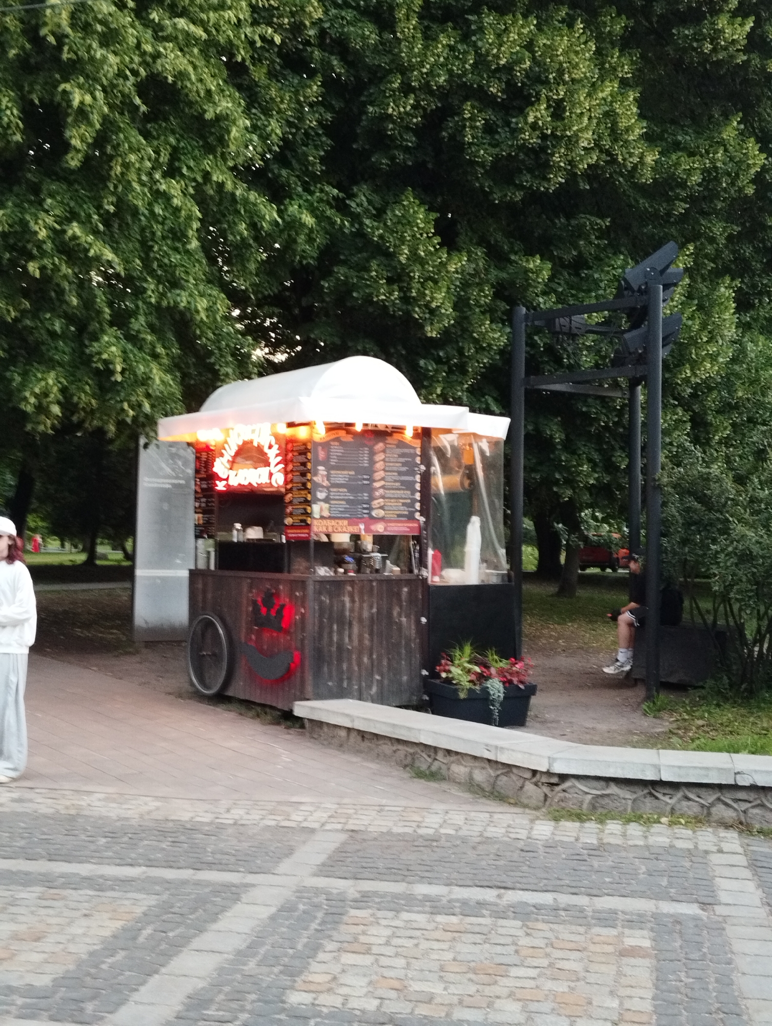 This image depicts a food cart situated outdoors in a park-like setting. The cart is constructed of dark wood with a white awning illuminated by red and orange lights. A menu is displayed on the front of the cart. In front of the cart, a person wearing a white hoodie and light-colored pants stands looking to the left. To the right of the cart, a person is seated on a dark structure, appearing to be a bench or seating area. The background features abundant green trees and a cobblestone walkway in the foreground. The scene suggests a casual outdoor dining or recreational area.