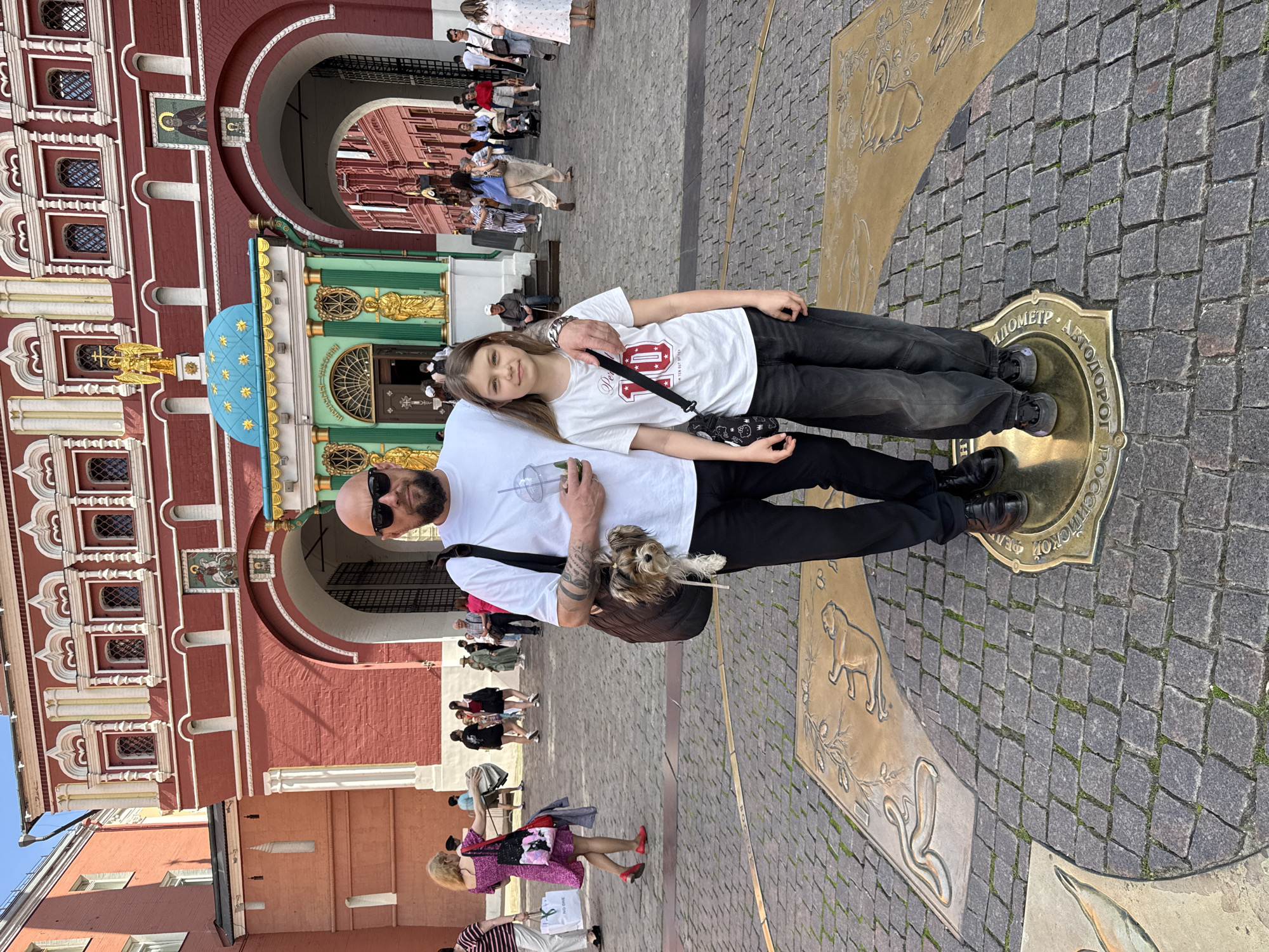 A man and a young girl pose for a photograph on a cobblestone square in Moscow, Russia. The man, with a beard and sunglasses, wears a white t-shirt and black pants, holding a clear cup. A small dog with light fur peeks out from a black bag slung over his shoulder. The girl, with long light brown hair, wears a white t-shirt with a red "UNI" design (possibly "07" or "01"), black jeans, and sneakers, holding a small black and white bag. She leans slightly into the man, who has his arm around her shoulder.
They stand on a large circular bronze plaque embedded in the ground, marked "КИЛОМЕТР • АВТОДОРОГ РОССИЙСКОЙ ФЕДЕРАЦИИ" (KILOMETER • MOTORWAYS OF THE RUSSIAN FEDERATION), which is Moscow's Kilometer Zero marker. Adjacent to it are other bronze plaques with engraved animal designs, including a lion-like creature, a snake-like creature, and a bird.
The background features the ornate architecture typical of Red Square. To the left is a red building with multiple windows framed by white decorative trim. Through a large arched gateway, another red building is visible. Centrally, behind the man and girl, stands the green and gold Iberian Chapel, topped with a blue dome adorned with golden stars and a golden eagle statue. A religious icon with Cyrillic text below it is above an archway near the chapel. Numerous other people are visible in the background, walking around the busy square. The scene is set on a bright, sunny day with clear skies, suggesting pleasant weather. The subjects appear relaxed and are likely posing for a commemorative photo at this landmark location.