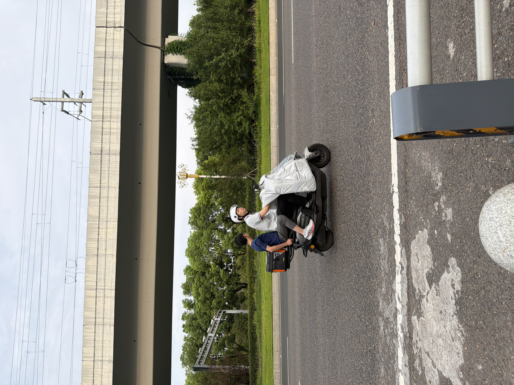 The image shows two people riding an electric scooter on an asphalt road in Jiangjin, China. The scooter has a black storage box on the back. The person in front is wearing a white helmet and a white shirt with red accents. The person in the back is wearing a blue t-shirt and gray pants. They are traveling under a large concrete overpass, possibly a highway or railway bridge. Behind the overpass, there are lush green trees and vegetation. Overhead, there are power lines and some infrastructure related to transportation. The sky is overcast. In the foreground, there is a white line marking the edge of the road and a portion of a grey and yellow barrier.