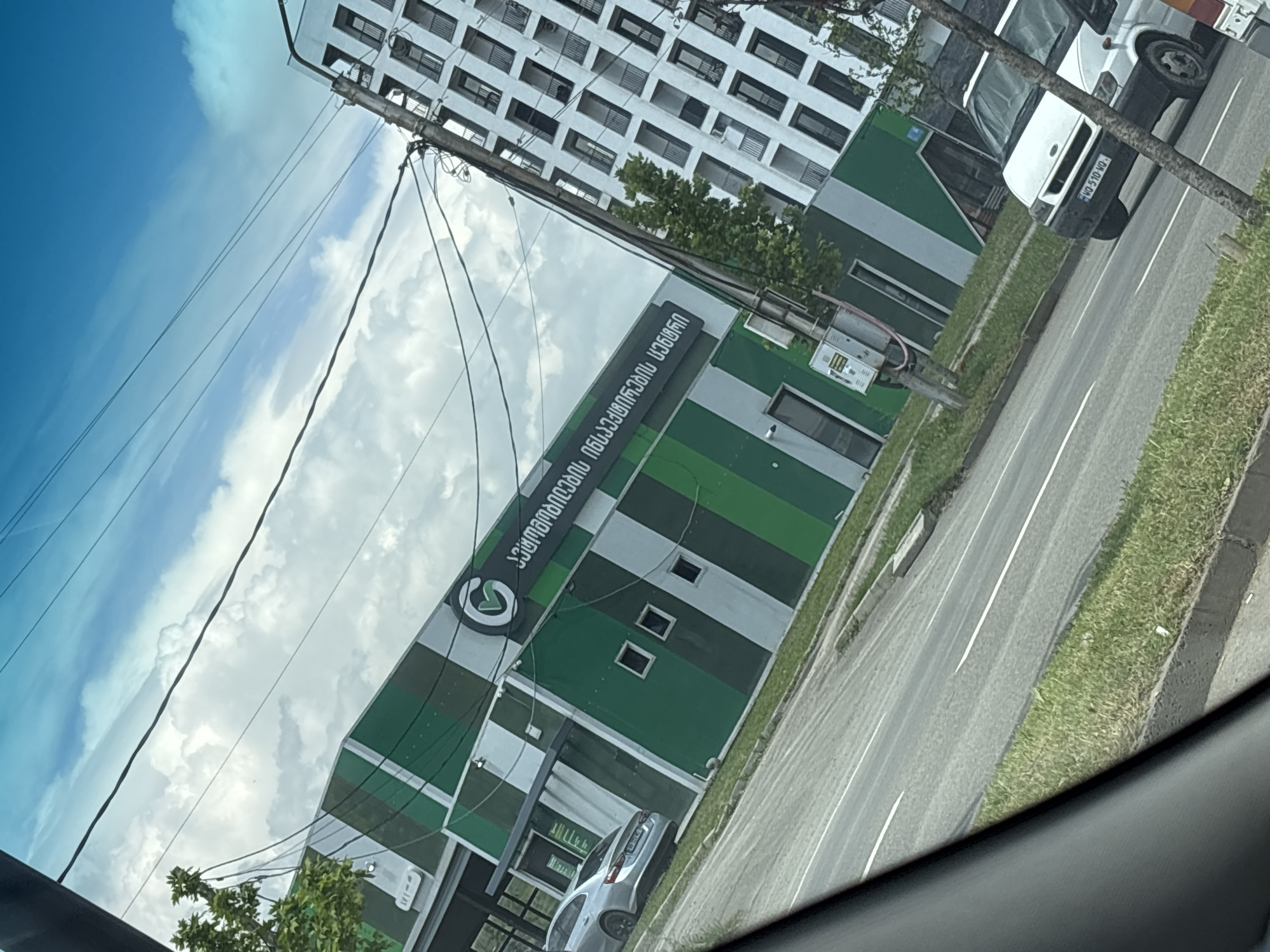 A slightly rotated low-angle shot depicts a street scene in Tbilisi, Georgia. A modern building with green and white horizontal stripes and a sign in Georgian script is visible. The sign includes a logo. A tall apartment building is visible in the background. A white van is parked on the side of the road. A silver car is parked near the striped building. The sky has a mix of blue and white clouds. Utility lines are strung overhead. A road and a grassy area are on the right side of the image. The image appears to be taken from inside a vehicle, as indicated by the interior trim visible at the bottom.