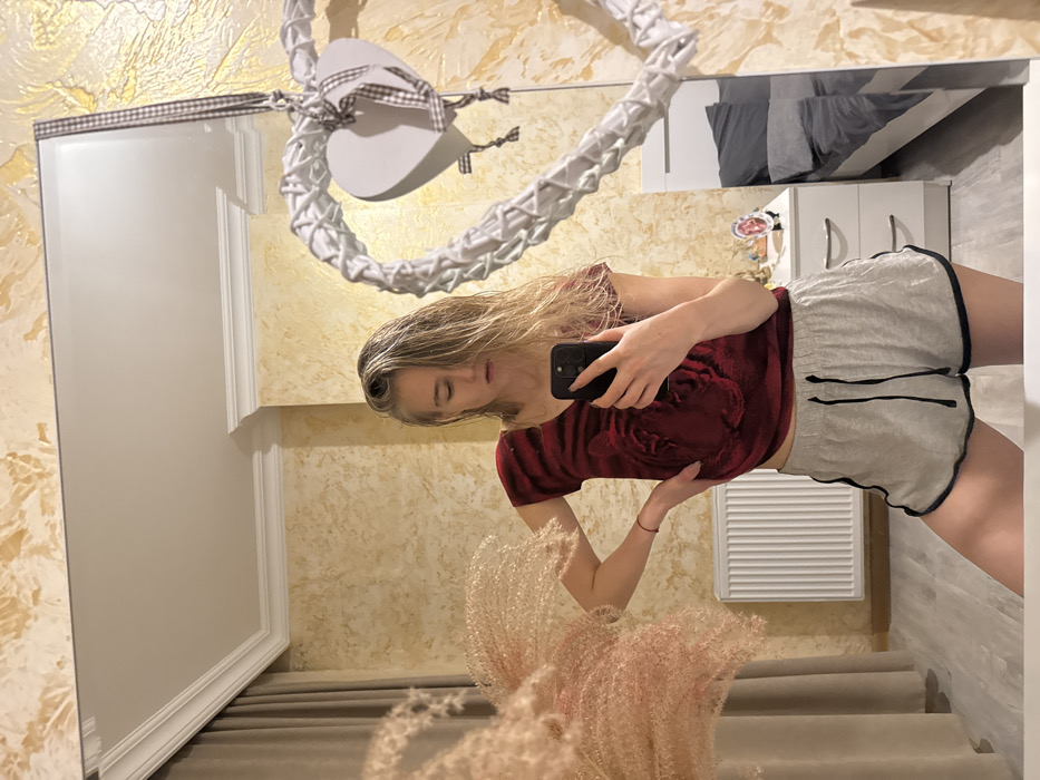 A mirror selfie captures a woman in a red top and grey shorts. She holds a phone in her right hand, reflecting a room with textured walls, a white dresser, and a bed. A white heart-shaped decoration hangs above, and decorative flowers are visible in the foreground. The mirror’s frame is partially visible, and a glimpse of stairs is caught in the reflection.