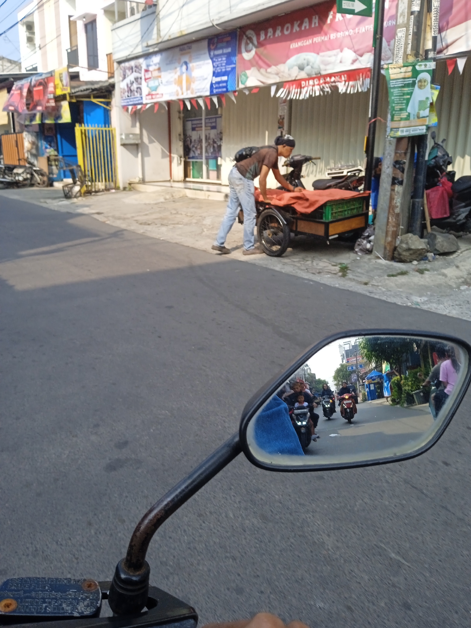 The image shows a street scene in Bekasi, Indonesia. In the foreground, a motorcycle's side mirror reflects a street with several motorbikes and people. One motorbike in the reflection has a child riding with an adult. On the right side of the reflection, a person in a pink shirt is visible. In the background, a man in jeans and a t-shirt is leaning over a three-wheeled cart loaded with an orange tarp and green crates. He is standing on the sidewalk next to a building with a sign that reads "BAROKAH FROZEN". The street appears to be paved with asphalt, and there are some plants and debris along the roadside. Buildings line the street, some with banners and advertisements. The overall scene depicts a typical day on a busy street in an Indonesian city.