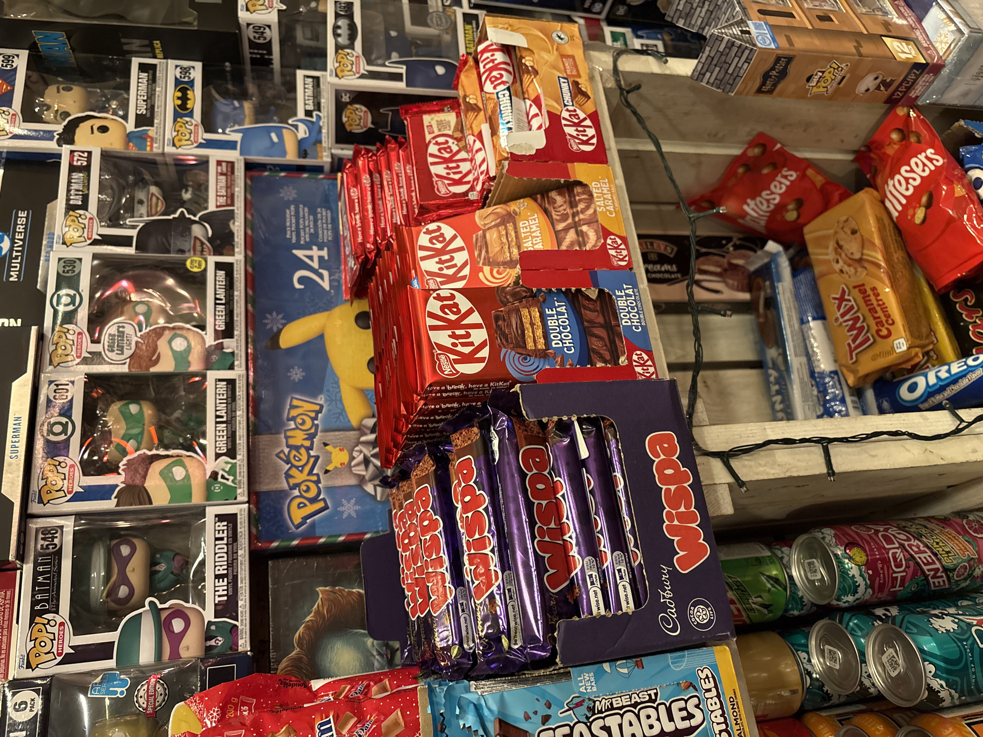 This is a photograph of a shelf stocked with an assortment of items, likely in a store setting. The products are a mix of toys and candies, densely arranged to maximize display space.
On the left are several Funko Pop! figures of DC Comics superheroes, including Superman, Batman, and Green Lantern, still in their original packaging.
The center of the image features a stack of KitKat chocolate bars in various flavors, such as Double Chocolate, Salted Caramel, and Peanut Butter Chunky. Adjacent to the KitKats is a box of Cadbury Wispa chocolate bars.
Behind and alongside the candies, there are bags of Reese's Pieces, Twix Caramel Centres, and Oreo. Beneath the candies are rows of various canned drinks.
Additional details include a Pokemon-themed item (possibly an advent calendar) and a MrBeast Feastables chocolate bar. The lighting suggests an indoor environment with artificial lighting. The overall impression is one of a cluttered but well-stocked retail display, likely in Saint Petersburg, Russia.