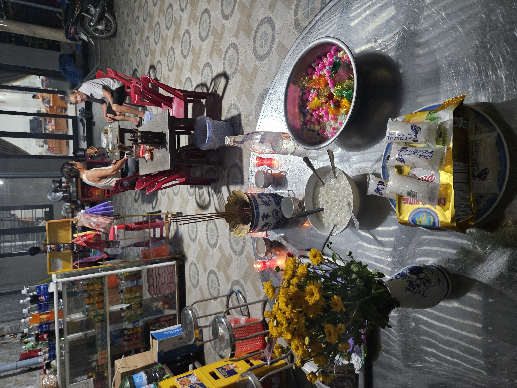 The image depicts an outdoor gathering or ceremony taking place in what appears to be the front yard or patio of a residence in Đà Nẵng, Vietnam. In the foreground, a metal table is laden with various items. A bouquet of yellow chrysanthemums in a blue and white patterned vase stands on the left. To the right of the flowers, there are two small white cups, a red tea light candle, and a ceramic incense burner with lit incense sticks. A bottle of clear liquid, likely water or alcohol, is positioned next to the cups. Further to the right, two bowls are visible: one containing white rice and another, a larger metal bowl, filled with a colorful assortment of candies and what appears to be rice. In the lower right corner, several packaged food items, some in yellow packaging, are stacked.
In the mid-ground, two men are seated at a rectangular table with red chairs. They are engaged in conversation or observation. The man on the left wears a white tank top, while the man on the right is in a light-colored short-sleeved shirt and dark shorts. The table they are at has chopsticks and other items, suggesting a meal or offering.
The background reveals a building with a large open doorway, through which a room with furniture and a television can be seen. A motorcycle is parked on the right side of the frame, partially obscuring a building wall. The ground is tiled with a circular pattern. A shelf unit filled with various bottles and containers of drinks and food is visible on the far left, suggesting a small shop or pantry area integrated into the outdoor space. The overall lighting suggests it is either early morning or late evening. The atmosphere appears to be one of a casual gathering or possibly a ritualistic observance.