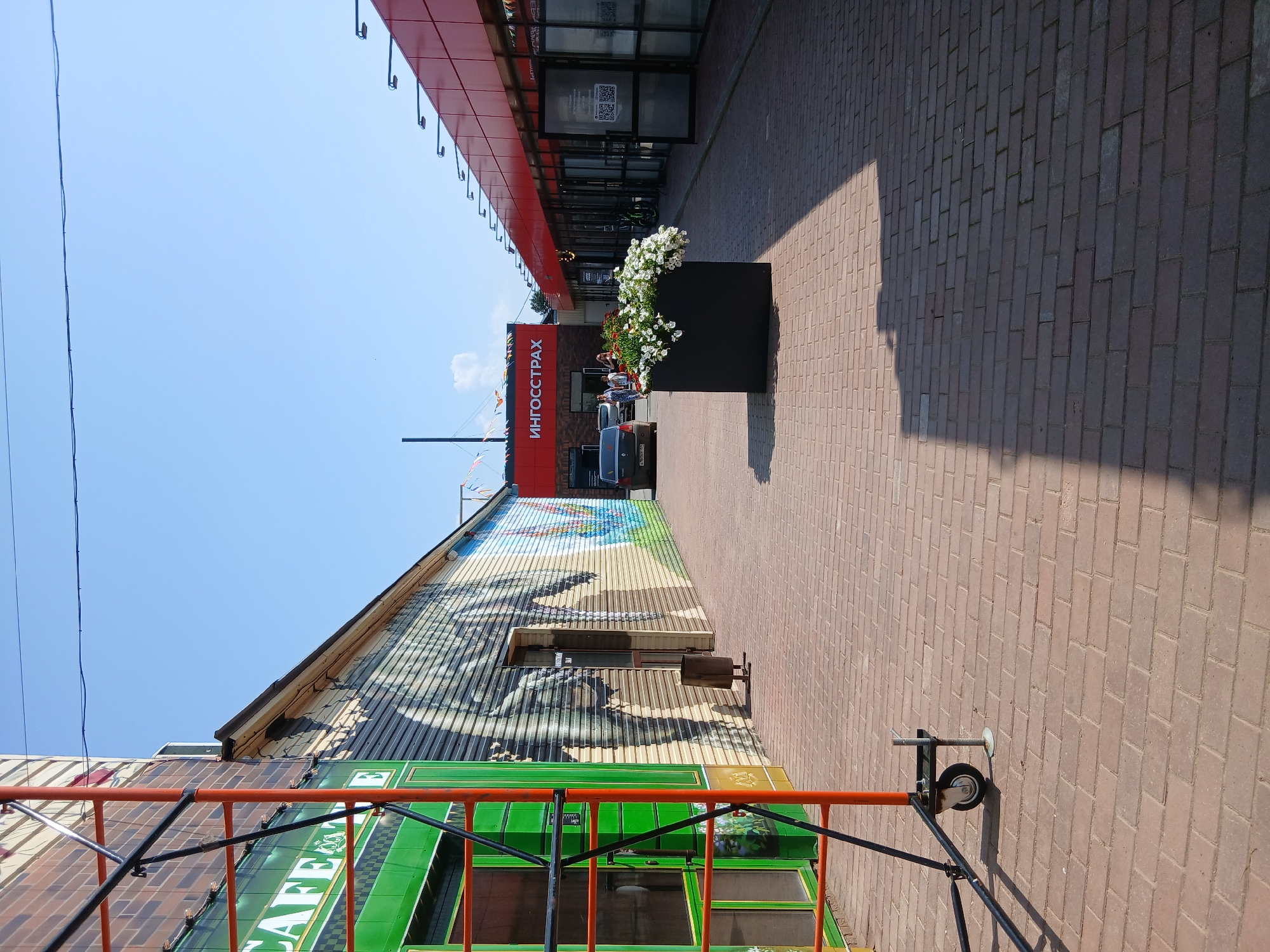 The image depicts an outdoor scene in the city of Osa, Russia, likely a commercial area. In the foreground, a brick-paved walkway is visible, with scaffolding to the left. The scaffolding has orange and black metal frames and a wheel. Adjacent to the scaffolding is a building with a green facade and "CAFE" signage. The building's wall features a large mural with a depiction of an animal, possibly an elephant, painted in shades of grey and brown, alongside a colorful landscape. Further down the walkway, there is a black planter box filled with white flowers.
To the right of the planter box, a red-colored building with multiple glass-fronted shop entrances is present. One of these entrances is ajar, revealing an interior. Above the shops, the red building has a series of horizontal light fixtures. Further to the left, a black car is parked, and a sign reading "ИНГОССТРАХ" (INGOSTRAKH) is prominently displayed on a building in the background. Several people are walking in the distance near the "ИНГОССТРАХ" building and the parked car. The sky is clear and blue, suggesting a sunny day. The scene appears to be a pedestrian area in front of shops and businesses.