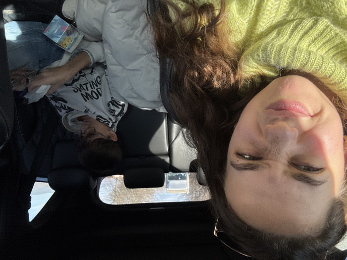 The image displays two individuals inside a vehicle in Banichan, Bulgaria. In the foreground, positioned on the left, a female with long, dark, wavy hair wears a lime green ribbed turtleneck sweater. Her face is directed towards the viewer, with a wrinkled nose and slightly puckered lips, and sunglasses are resting on her head. Sunlight illuminates her face from the right. In the background, seated in the right rear passenger seat, is a younger male wearing glasses, a white hooded sweatshirt with dark lettering partially visible as "SKATING MOD," and blue jeans. He is looking down, engaged with a small white rectangular object and a blue and teal box, possibly card packaging, held in his hands. A white puffy jacket or blanket is visible beside him. The vehicle's interior consists of dark seats and headrests. Through the rear window, a bright blue sky, bare trees, and distant hills are visible, indicating an outdoor setting.