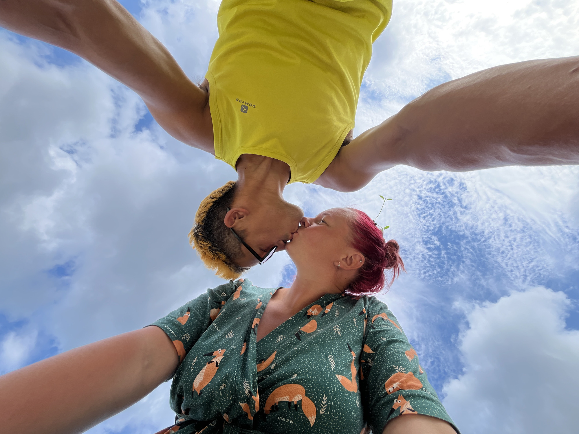 A low-angle shot captures two individuals kissing under a cloudy blue sky. The person on the left, wearing a green dress with an orange fox pattern, has pink hair pulled up in a bun. A small green sprout is visible near their head. The person on the right, wearing a yellow tank top with a "DOMYOS" logo, has short, dyed blond hair and is wearing glasses. Their arms are raised, with the person on the right's leg extended upward, creating a sense of connection between them and the sky. The background is dominated by a bright blue sky with scattered white clouds. No specific location in Chaikovsky, Russia, can be identified from the image.