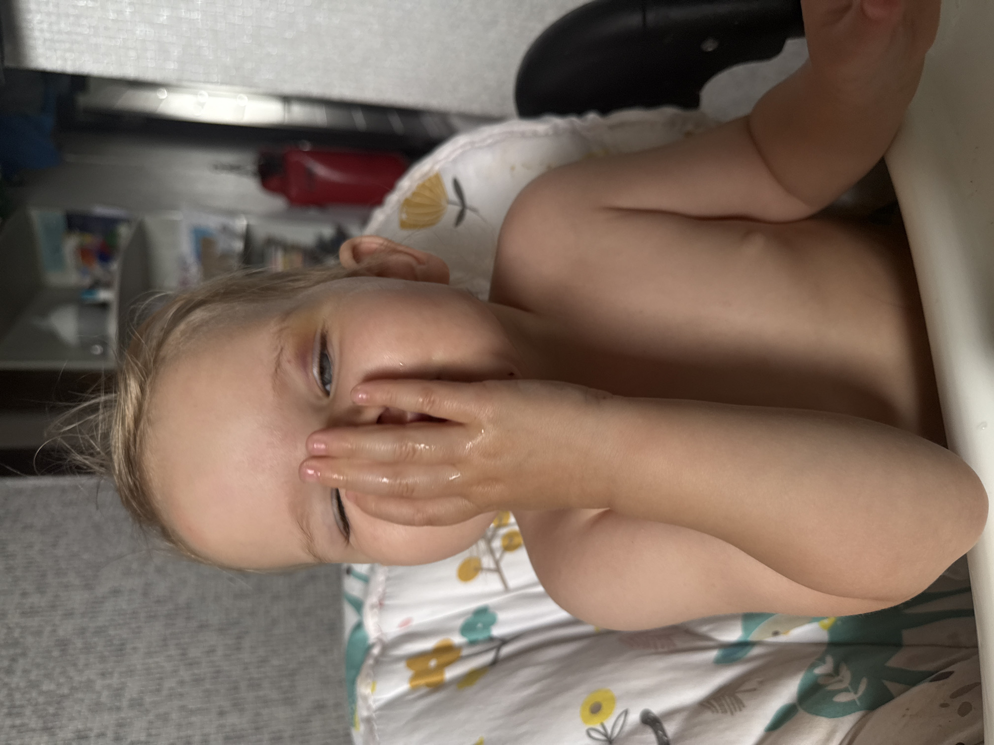 This is a close-up, vertically oriented media file of a young child. The child has blonde hair and is partly covering their face with their hand. Their eyes are visible, and they appear to be looking towards the viewer. The child is wearing a patterned bib or blanket. The background is blurred but suggests an indoor setting with furniture and possibly shelves with items. There is no visible text. The lighting appears to be natural or indoor artificial light. The image is likely taken in Moscow, Russia, given the provided location context.