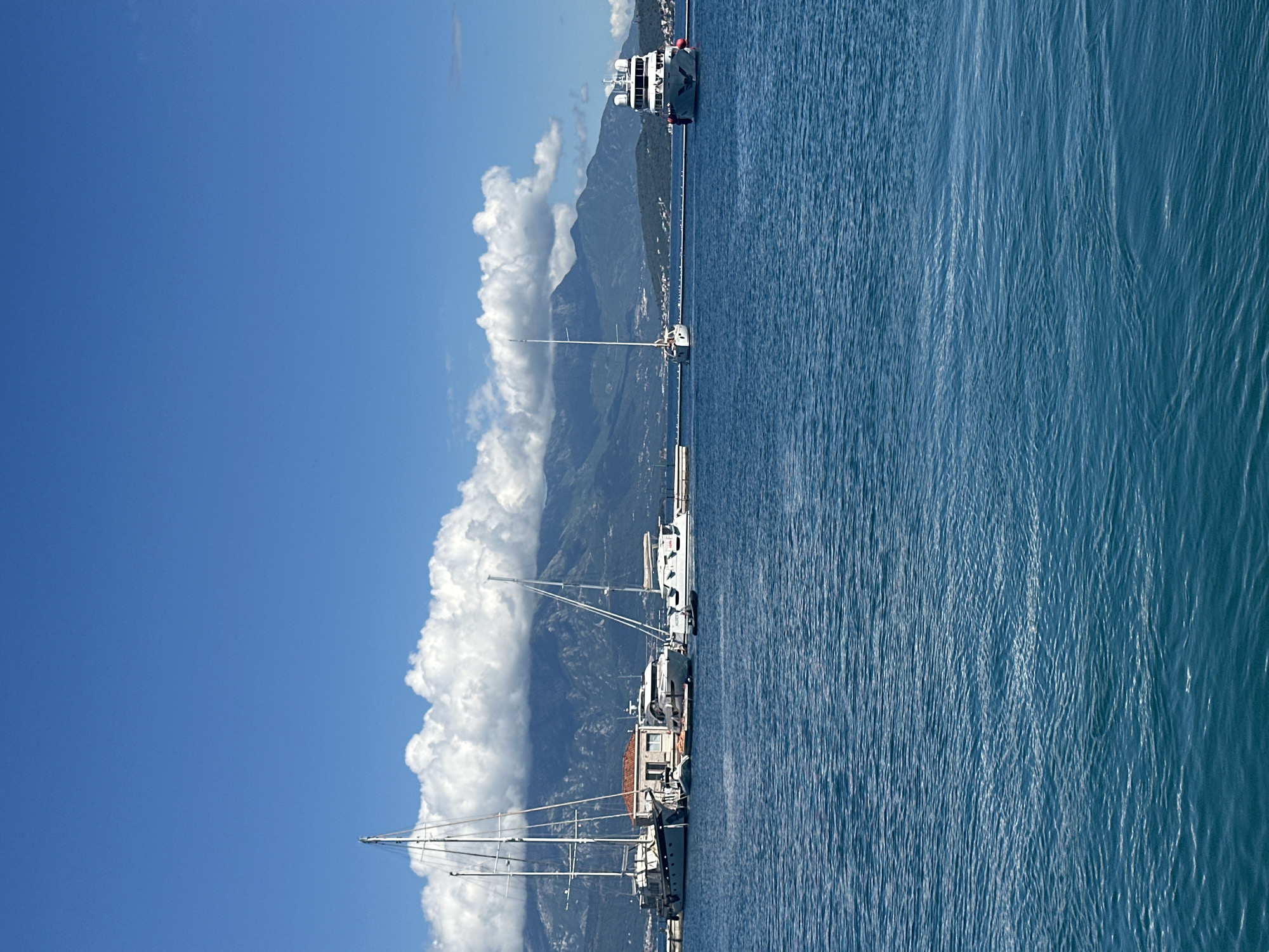 This image presents a serene and picturesque coastal landscape, likely depicting a bay or fjord near Herceg Novi, Montenegro, as suggested by the location context.
The scene is dominated by a clear, vibrant blue sky filling the upper portion of the frame, indicating bright, sunny weather. Below the sky, a dramatic line of majestic mountains rises from the water, their slopes covered in green foliage. A striking feature is a long, continuous bank of fluffy white cumulus clouds clinging to and partially obscuring the mountain peaks, creating a soft, ethereal contrast with the rugged terrain.
In the midground, along the base of the mountains, a series of boats are moored or docked. These include several sailboats with their tall masts and rigging silhouetted against the clouds and sky. Further along the dock, a large, prominent white motor yacht or ship with multiple decks is visible, featuring a distinctive red life raft on its stern. Other smaller vessels are also present along the shoreline.
The foreground and much of the midground are covered by calm, deep blue water, showing gentle ripples and reflections that suggest a tranquil environment. The overall impression is one of peaceful maritime activity set against a stunning natural backdrop of mountains and a clear sky. The time of day appears to be bright daylight, possibly mid-day, given the strong illumination. There is no visible text in the image.
