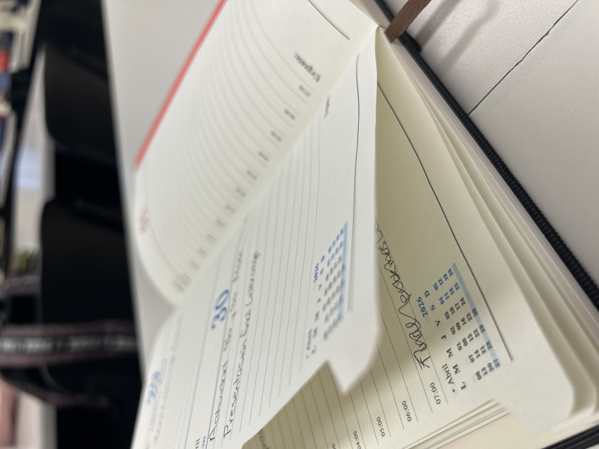 An open planner or diary is displayed from an elevated perspective, resting on a surface in an indoor setting, possibly an office, with blurred dark furniture in the background. The planner's pages feature both printed and handwritten entries, indicating planning or record-keeping.
Visible printed text includes a calendar grid for "Abril" (April) 2026, showing dates from 01 to 30, and days of the week labeled "L M M J V S D". Time slots "04:00", "05:00", "06:00", and "07:00" are printed on the daily planning pages. The date "30" is prominently featured.
Handwritten notes on the page for the 30th read "Actividad 96" and "Presentación fund Liversage". Another handwritten phrase, "Final probkibas" (possibly "Final pruebas"), is visible. Other partially legible printed words include "Logistics" and "Urgenter" (likely "Urgente"). The number "245" is also present on one of the pages. The foreground is in focus, while the background and some distant pages are blurred due to shallow depth of field.