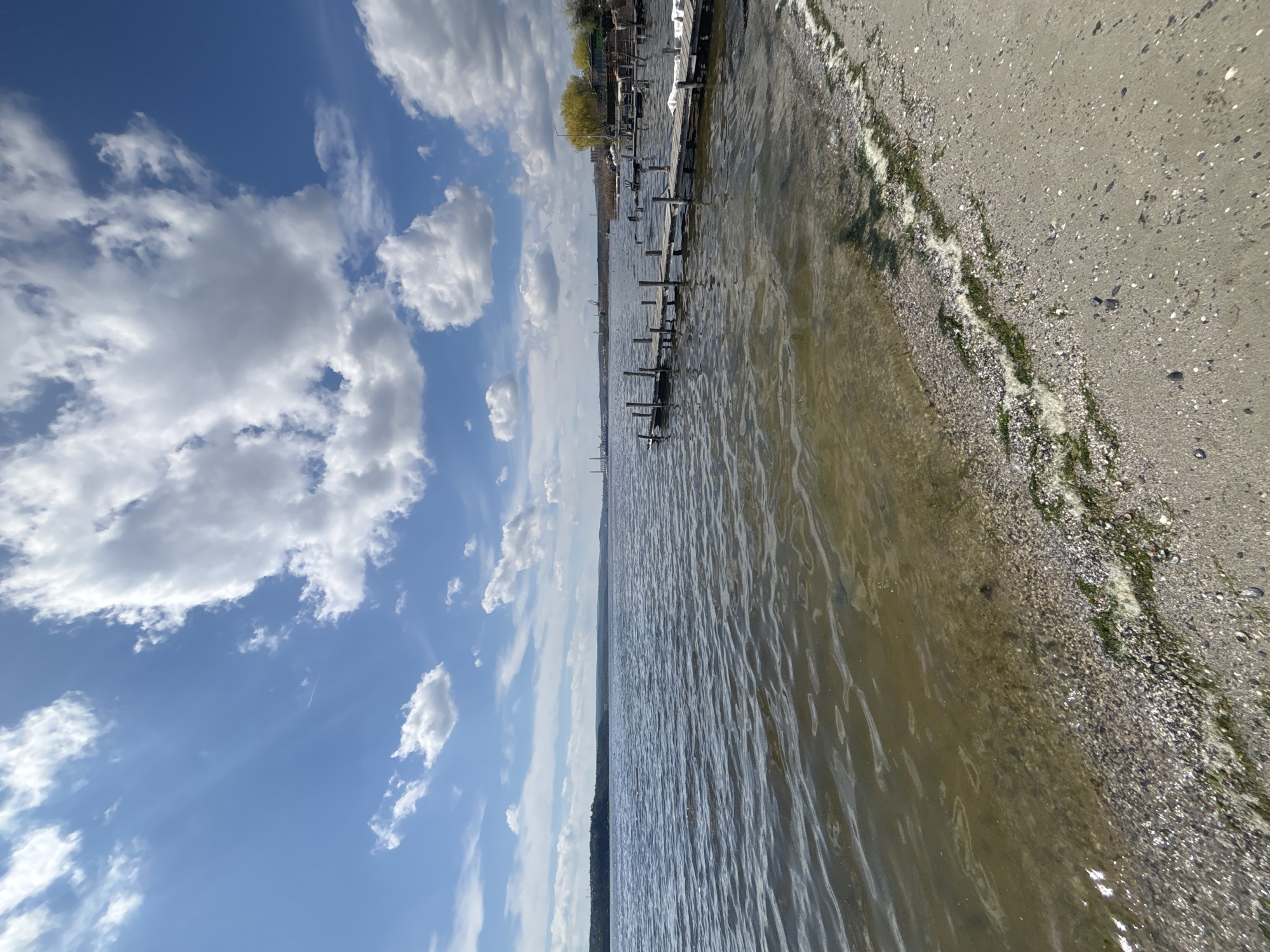 The image depicts a landscape view in Казашко, Bulgaria. The scene features a body of water, possibly a lake or the sea, extending towards the horizon. Along the right edge of the frame, there is a sandy beach with seaweed or algae scattered along the shoreline. An old, weathered wooden pier or dock is visible extending into the water. The sky occupies the upper left portion of the image, filled with cumulus clouds scattered against a blue backdrop. The lighting suggests it is daytime.