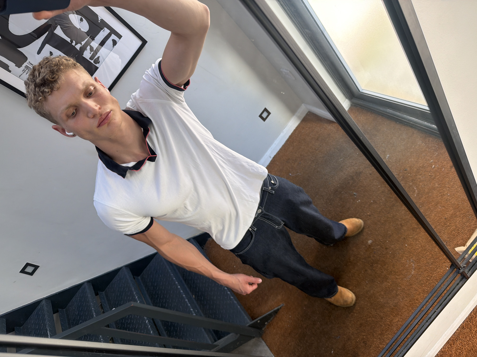 A fair-skinned man with blond curly hair and a white polo shirt is taking a selfie in what appears to be a modern apartment hallway in London, United Kingdom. He is wearing dark denim jeans and tan suede boots. The background shows a black metal staircase, a white wall with a small square black decoration, and a large framed black and white poster. Sunlight streams in from a large window on the right side of the image. A reflection of the man and his surroundings is visible in the glass door.