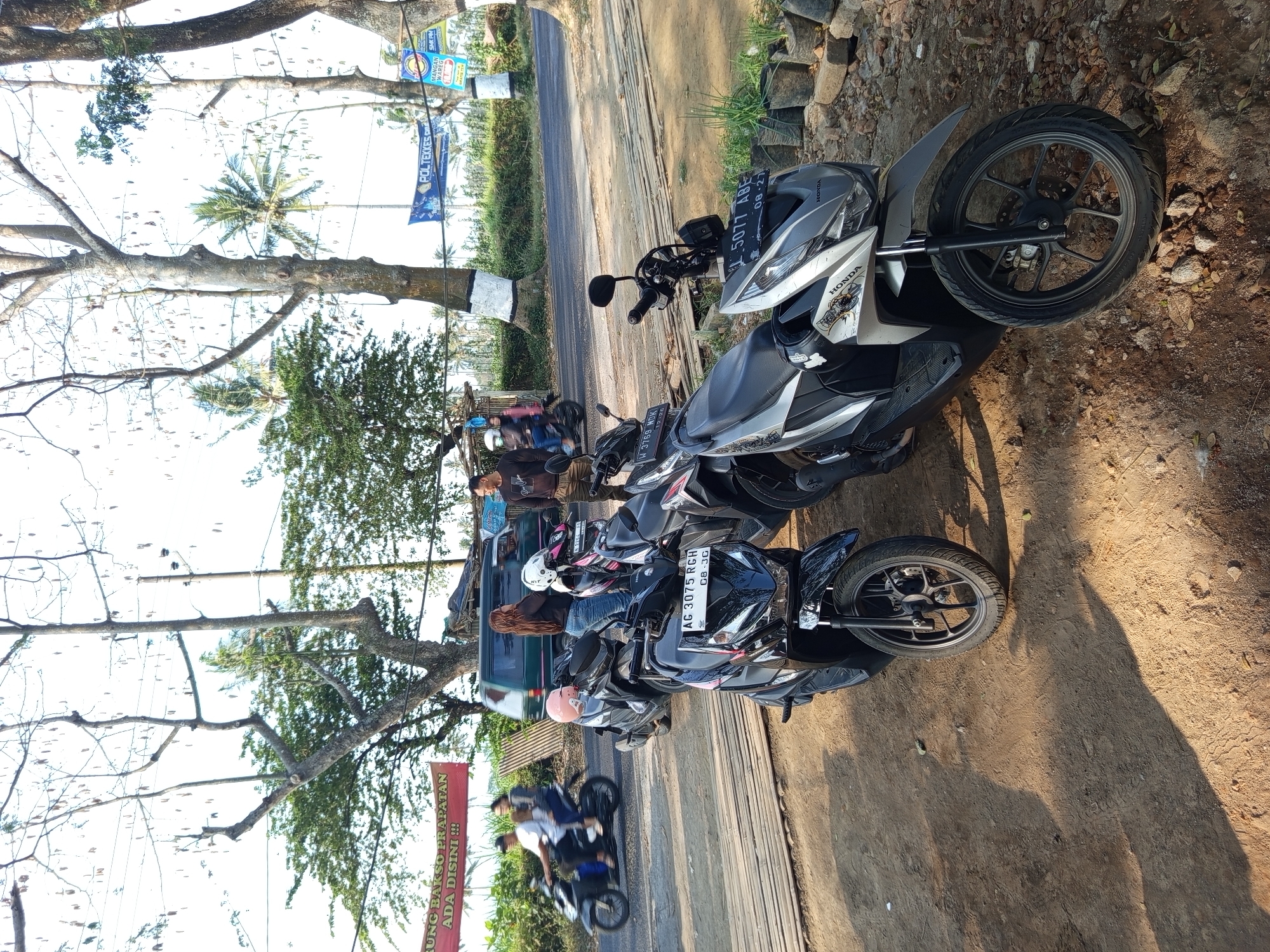 The image depicts a street scene in Bokor, Indonesia, with several motorcycles parked on a dirt and bamboo-planked area adjacent to a road. In the foreground, two automatic scooters are prominently displayed. The scooter on the left is black with pink accents, and its license plate reads "AG 3075 RGH". The scooter on the right is silver and black, with the license plate "L 5077 ABE". A third black scooter is partially visible behind these. In the background, a green van is parked on the asphalt road. A group of people are standing near the van, including a woman with long, dark hair and two men. Another person is riding a black motorcycle on the road, with a passenger behind them. Trees with bare branches and palm trees are scattered throughout the scene, with some vegetation and electrical poles visible. Two banners are hanging from poles in the background, one appears to have blue and white elements and the other is red with white text. The ground is a mixture of dirt, small stones, and scattered dry leaves. The lighting suggests it is daytime, with shadows cast on the ground.