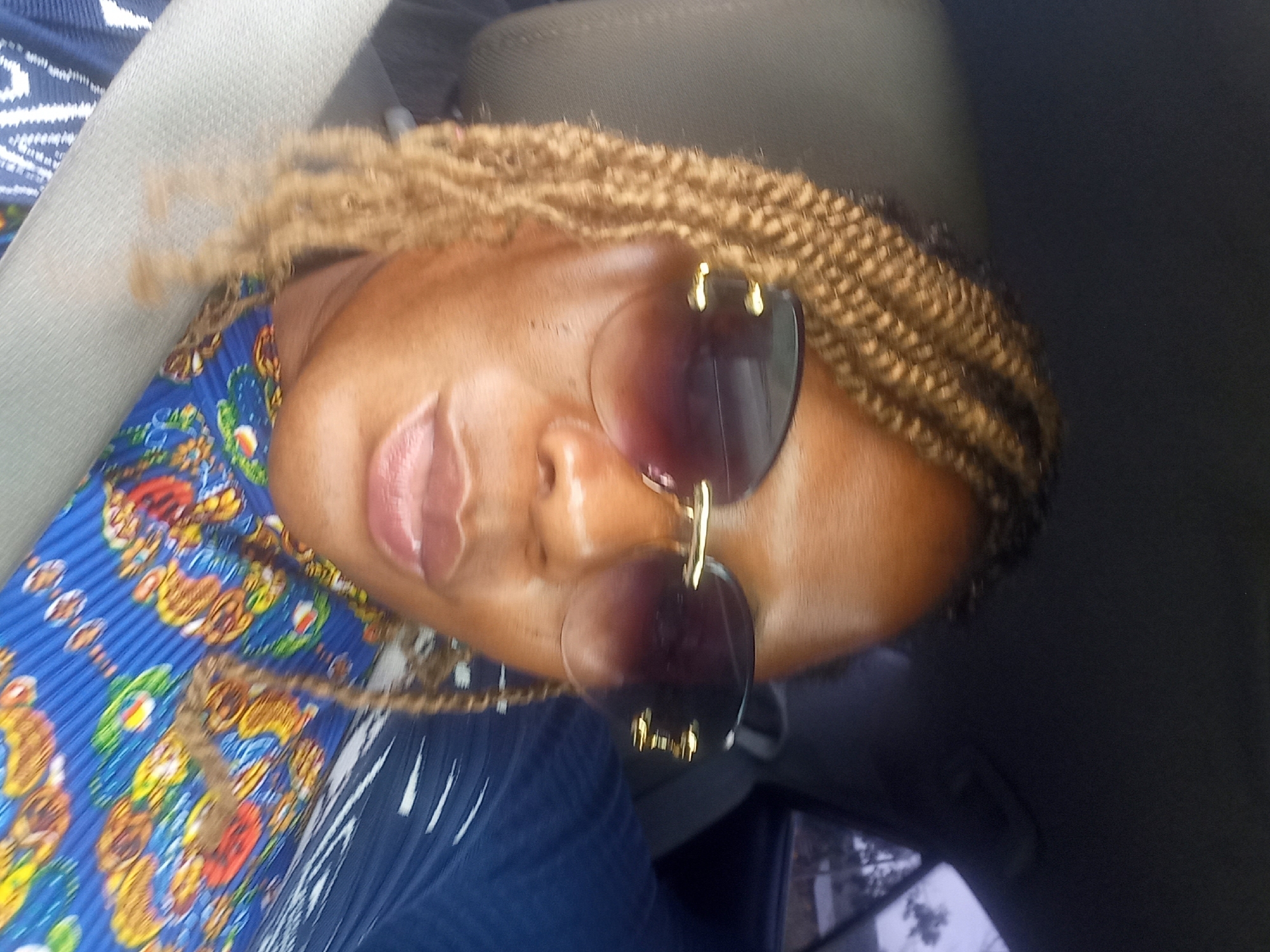 A medium shot captures a woman from the chest up, seated in the passenger seat of a vehicle. She is wearing large, dark-tinted sunglasses with gold accents and a vibrant blue top with a colorful, intricate pattern. Her braided, light brown hair frames her face. The background shows the interior of the car, including a seatbelt, and a glimpse of the outdoor environment through the rear window, suggesting the vehicle is in motion. The scene is likely Mangu, Plateau State, Nigeria, based on the reference to the city.