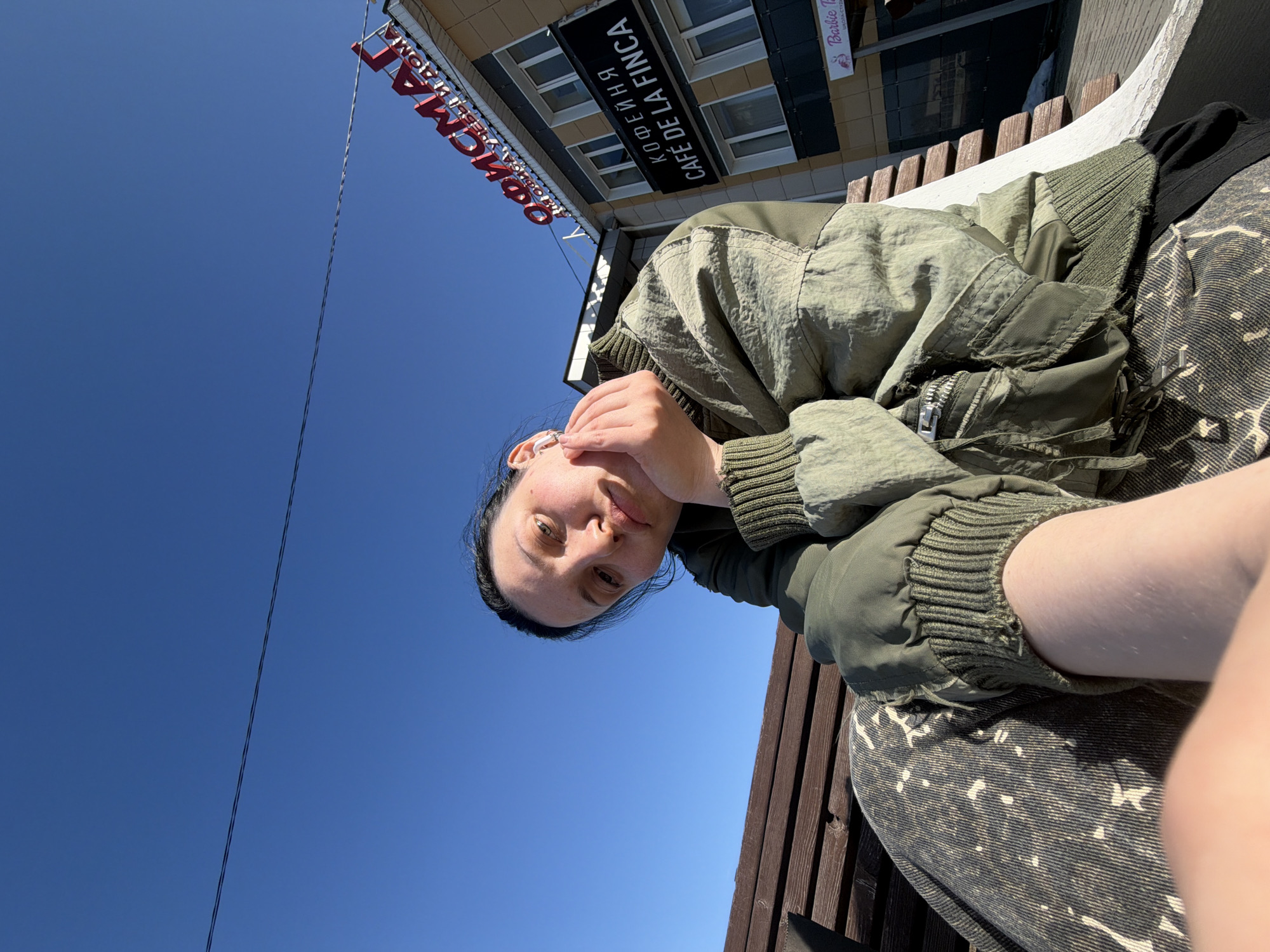 An individual, identified as female, is seated on a brown slatted wooden bench on ul. Lenina, 53, Chaykovsky, Perm Krai, Russia. She is positioned slightly to the left, facing directly towards the camera with her head tilted and her right hand supporting her chin. She wears an olive-green bomber jacket with ribbed cuffs and collar, and dark mottled pants. White wireless earbuds are visible in both ears. The background features a two-story commercial building with light-colored walls and multiple windows. The building displays prominent red Cyrillic lettering for "ОФИСМАГ" (OFISMAG) and a black sign with "КОФЕЙНЯ" (COFFEE SHOP) and "CAFÉ DE LA FINCA" in white text. Below "CAFÉ DE LA FINCA", a smaller sign indicates "Barbie T Школа-студия" (School-studio). The sky above is clear and bright blue, intersected by a single dark utility wire running horizontally across the upper frame. A small patch of snow is visible on the ground to the right of the bench. The scene is illuminated by strong sunlight.