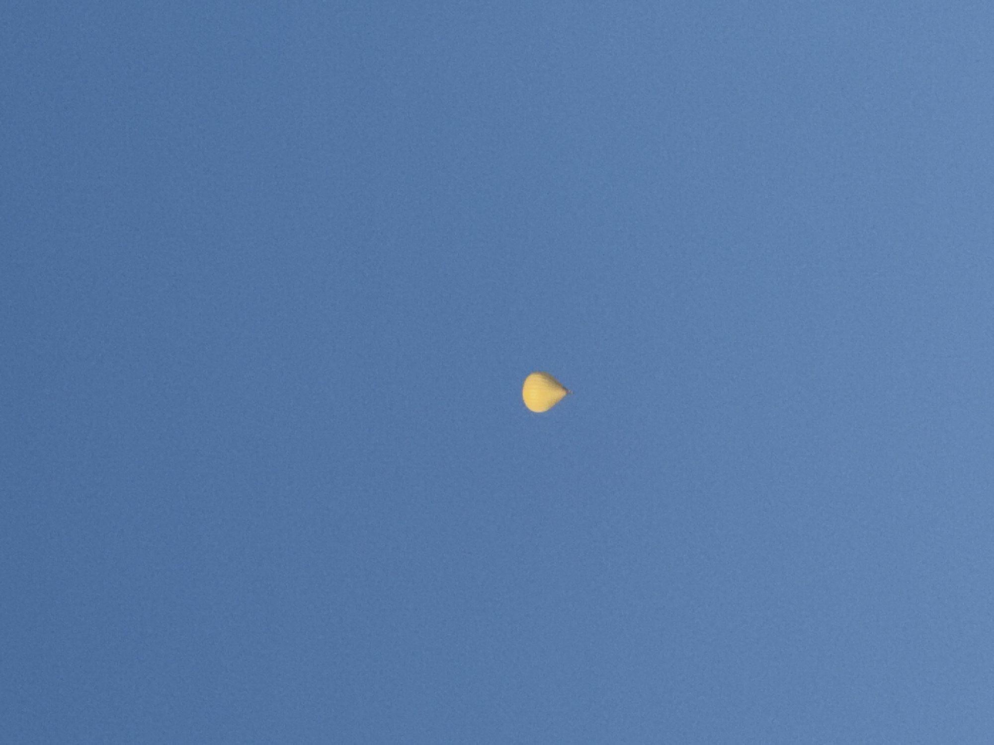 A yellow hot air balloon is visible in the bright blue sky. The balloon is small in the frame and appears to be at a significant altitude. The background is a clear, cloudless sky. The image does not contain any discernible landmarks or features that would indicate a specific location within Rimsting, Germany.