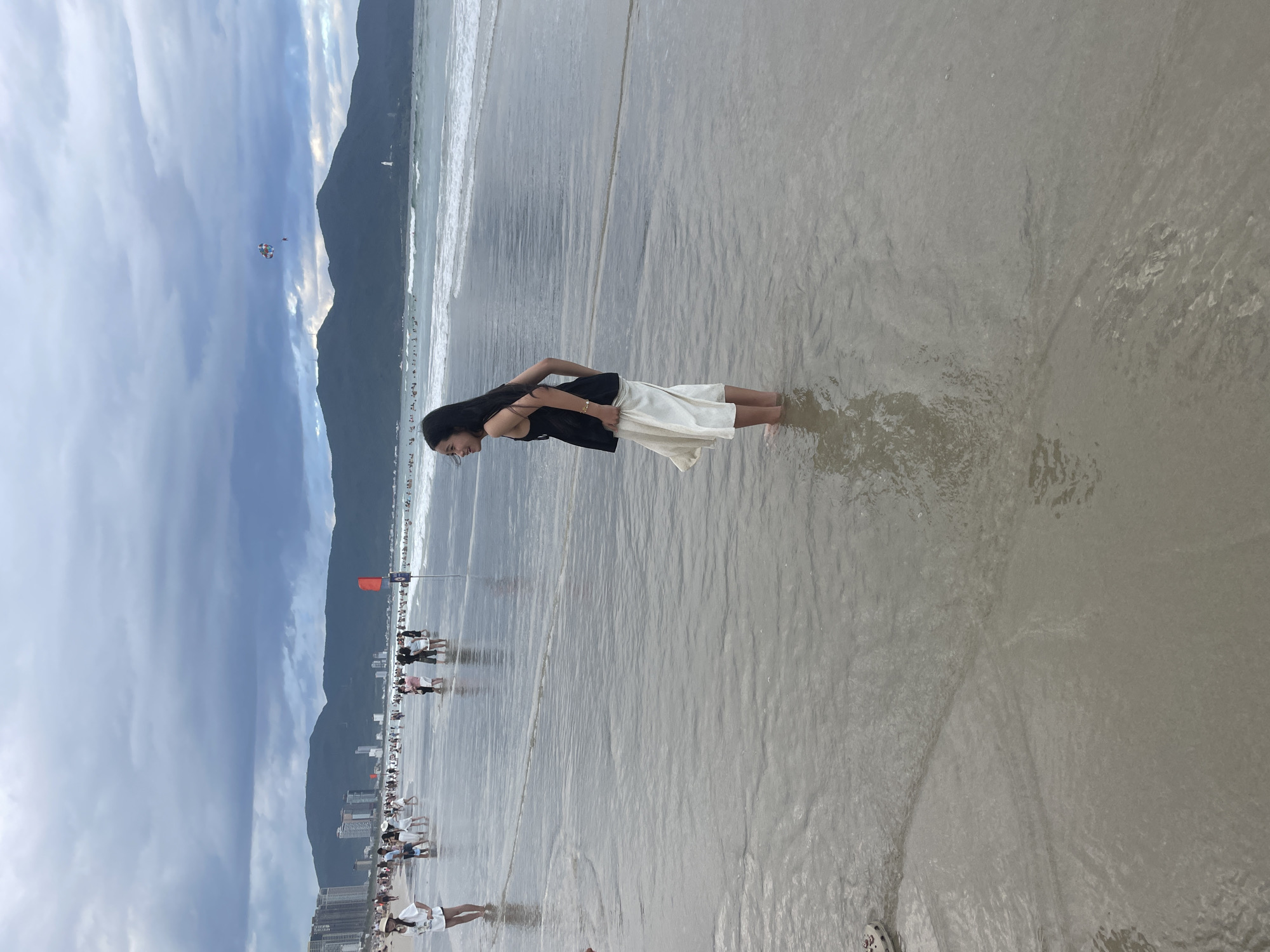 The image depicts a bustling beach scene in Da Nang, Vietnam, under a cloudy sky. In the foreground, a young woman with long dark hair stands ankle-deep in the shallow water, wearing a black sleeveless top and a flowing white skirt. She is facing away from the camera, looking towards the left of the frame. The beach itself is populated with numerous people, some wading in the water, others walking along the shore. In the background, a line of tall buildings and mountains forms the cityscape, with waves gently rolling in from the ocean. A parasailer is visible in the sky, adding to the recreational atmosphere. The water is calm and reflects the overcast sky.