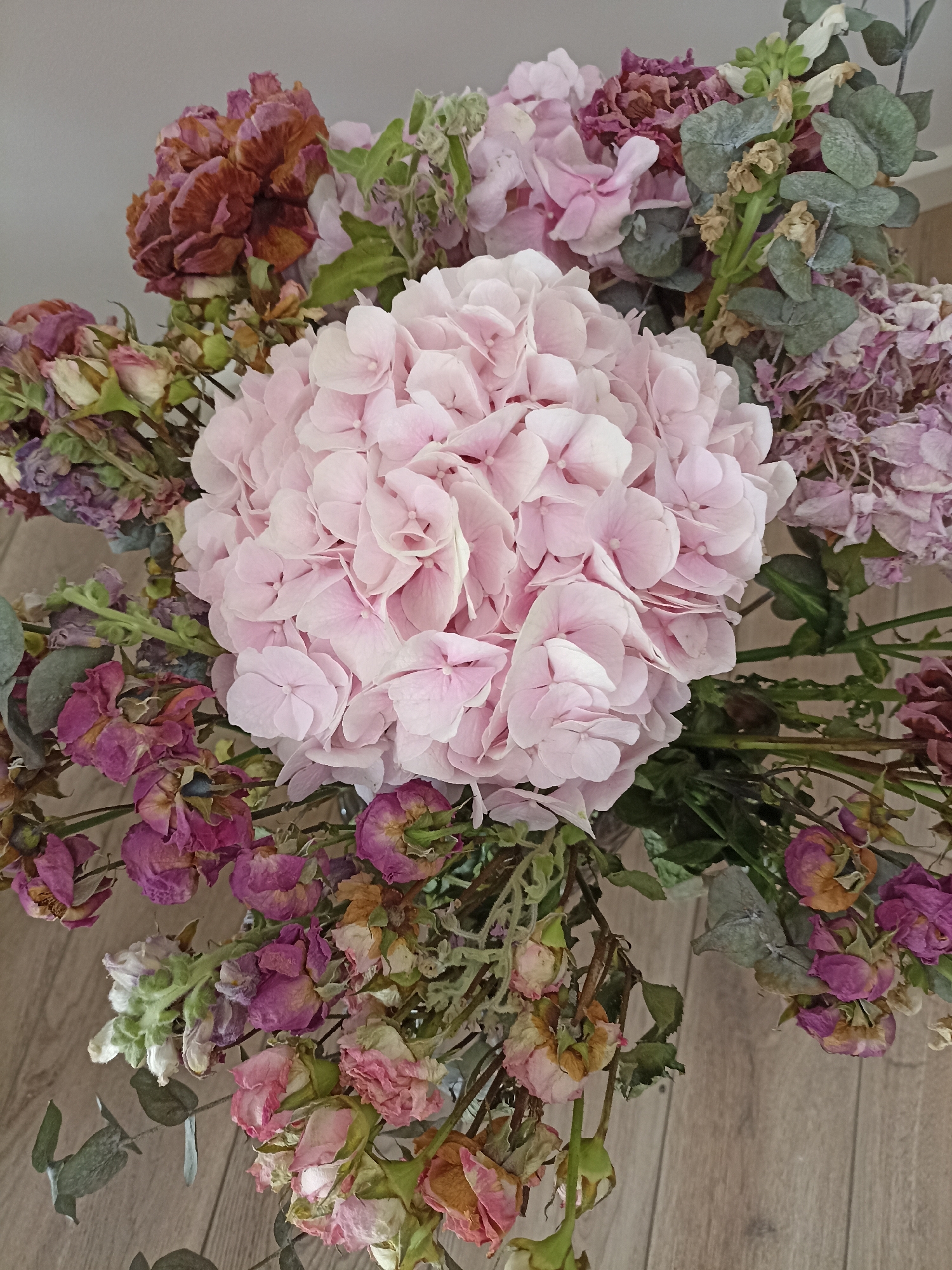 A high-angle shot captures a bouquet of dried flowers, predominantly pink hydrangeas and smaller, withered roses in shades of pink and purple. The flowers are arranged in a vase, though the vase itself is not fully visible. The bouquet is placed on a light brown wooden surface, likely a table or floor, with planks running horizontally. The background is a plain, neutral gray wall. The lighting is soft and diffused, casting no harsh shadows. The overall impression is one of gentle decay and preserved beauty. The scene is indoors, likely a home or studio, in an unspecified location.