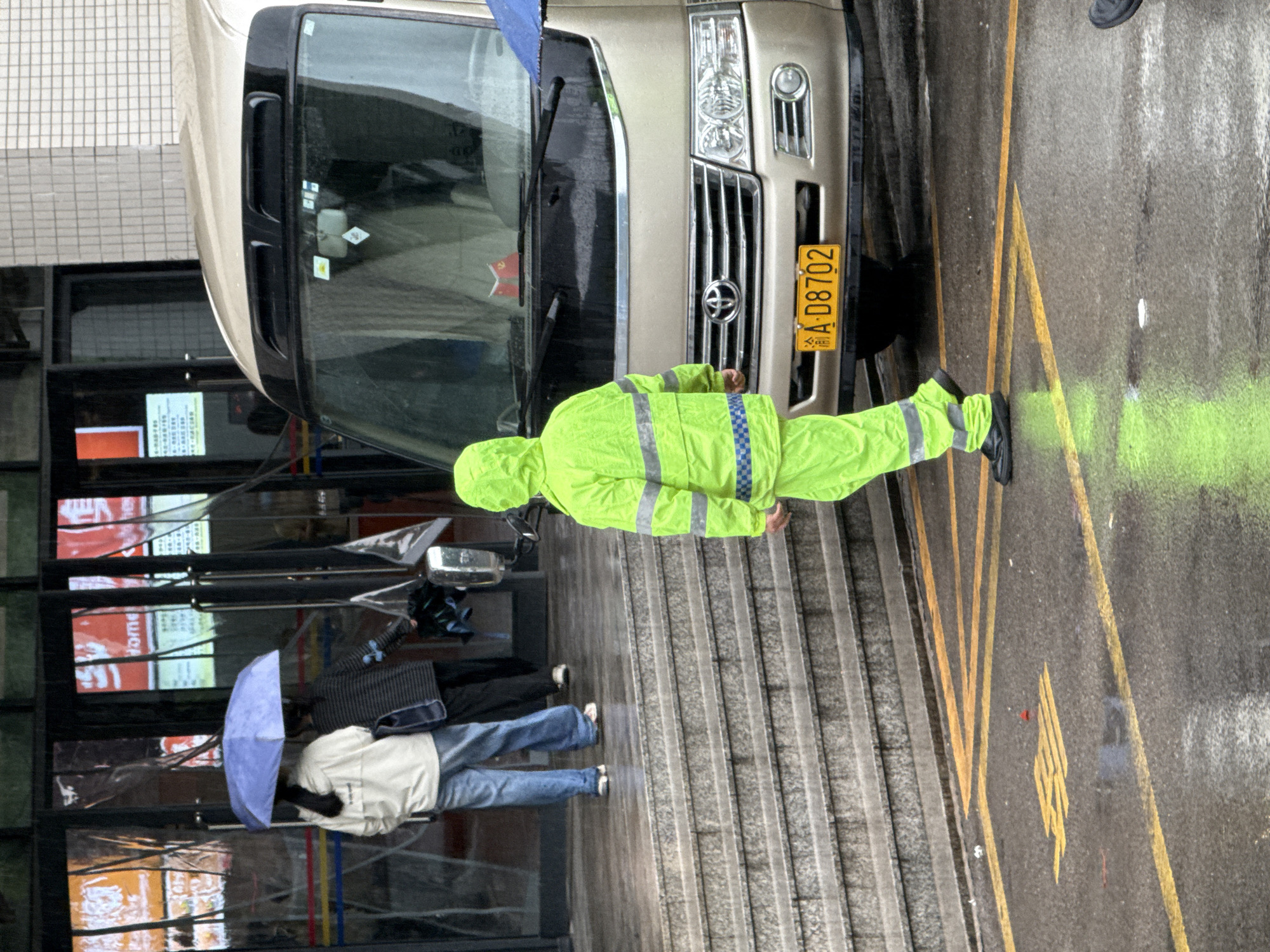 This image captures a wet, urban street scene, likely during or after a rain shower, as indicated by the damp ground and the presence of an umbrella. The setting is a street or public area alongside buildings.
In the foreground, a person dressed in a bright, neon green hooded reflective suit, detailed with blue and silver striped patterns, stands near the front of a gold-colored van. The person is facing away from the camera, towards the van's grille. The van, possibly a commercial or passenger vehicle, has a chrome front grille and headlights. A yellow Chinese license plate, reading "云 A D8702", is visible on its front bumper.
Further back and to the left, two individuals are seen. One person, wearing a light-colored jacket and blue jeans, holds a light blue umbrella, sheltering themselves and another person mostly obscured behind them. They are standing near a building with large glass windows that reflect colorful advertisements containing indiscernible text, and a tiled building facade is visible above the van.
The street surface is dark and wet, marked with yellow painted lines, including double solid lines and an arrow-like symbol. A distinct bright green spray-painted mark is also visible on the asphalt to the right of the person in the reflective suit. The general activity appears to be people navigating the street in rainy conditions. While the provided context states Jiangjin, China (Chongqing), the license plate "云" (Yun) typically denotes Yunnan province.