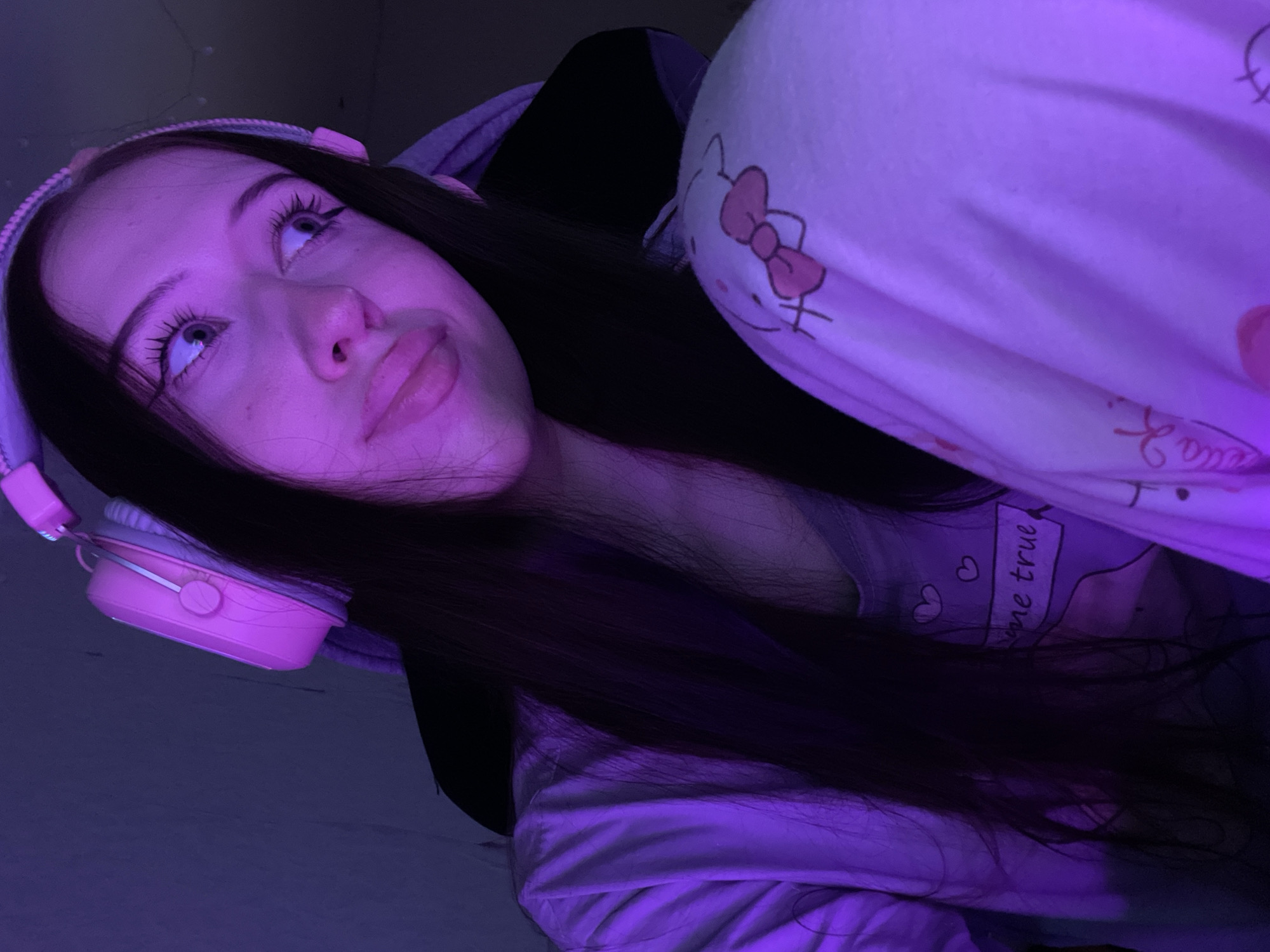 This is a close-up, low-angle photograph of a young Caucasian female with long, dark hair, wearing pink headphones and light-colored pajamas. The image is bathed in purple light, creating a dimly lit, atmospheric setting. Her eyes are open and looking to the right, and her lips are slightly parted. The pajamas have a cartoon print of Hello Kitty and other graphics. The background is dark and appears to be indoors, with a hint of a chair visible behind her. No specific location in Votkinsk, Russia can be identified from this image.