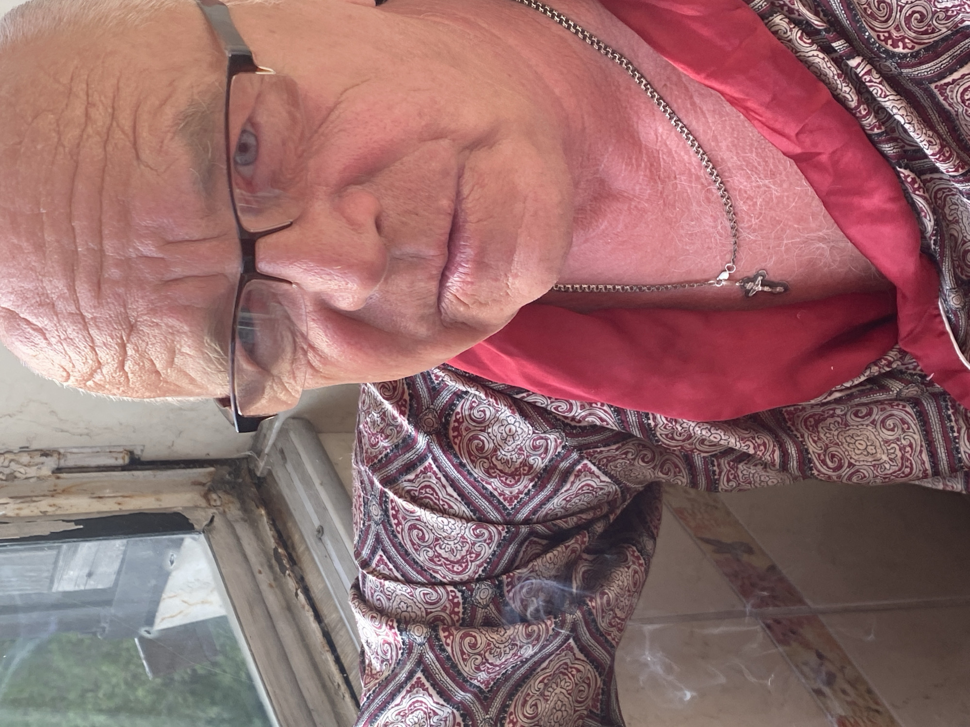 This close-up portrait, oriented sideways, features an older individual, likely male, with fair skin and prominent facial wrinkles, indicative of age. He has short, light-colored hair and wears dark-framed glasses, through which his light blue eyes are visible. His expression is neutral to slightly content. Around his neck, he wears a silver-colored chain necklace with a small, stylized cross pendant resting on his upper chest.
He is dressed in a richly patterned garment, possibly a robe or jacket, adorned with intricate maroon, off-white, and dark paisley-like designs. Beneath or as part of this, a plain red fabric is visible. A wisp of smoke, suggesting the act of smoking, rises near the lower edge of his patterned sleeve.
The setting appears to be indoors, adjacent to an old, weathered window frame with visible peeling paint, seen in the lower left corner. Through the window glass, green foliage and what appear to be outdoor wooden structures are faintly discernible, indicating a daytime setting. No visible text is present. The scene is set in Novokuznetsk, Russia.