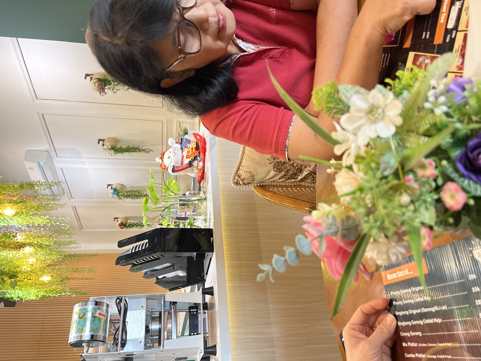The image depicts a woman in a red shirt sitting at a wooden table, looking down at a menu. She is wearing glasses and has dark hair. In the foreground, a colorful bouquet of artificial flowers is placed on the table. To her left, a hand is visible, also pointing at the menu. In the background, a counter area is visible with a cup sealing machine and a black cup holder. The wall behind the counter is decorated with hanging plants and a white air conditioner. A Maneki-neko (beckoning cat) figurine is also present on the counter. The overall environment appears to be a cafe or restaurant.