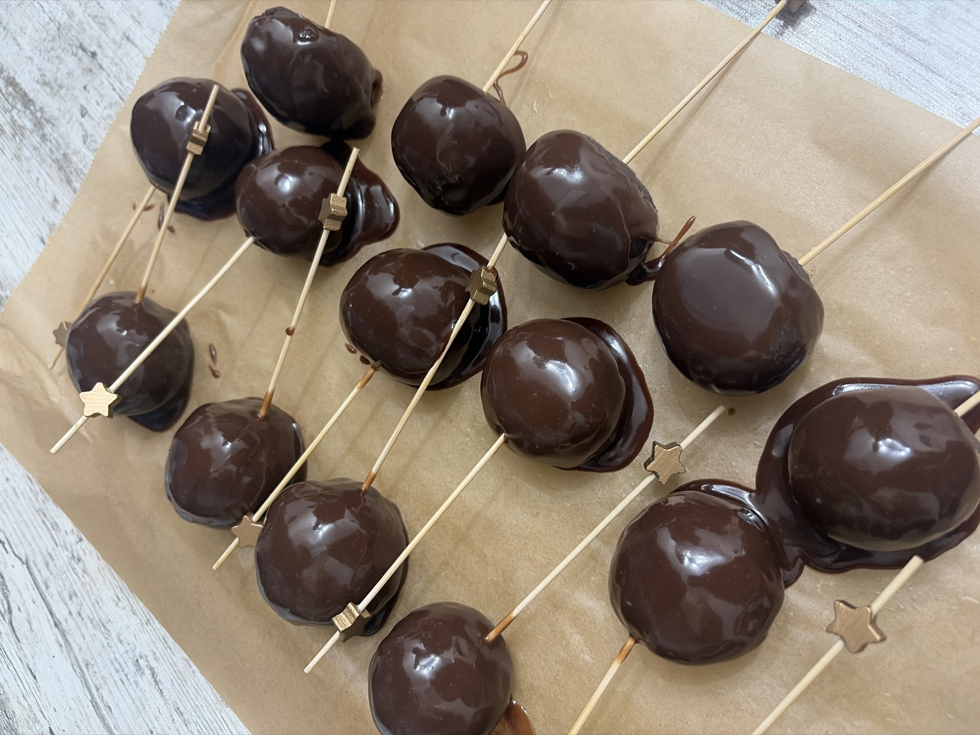 The image shows a close-up of chocolate-covered cherries on sticks, arranged on a light brown piece of parchment paper. The cherries have a dark, glossy chocolate coating. The sticks are wooden skewers with small gold star-shaped ornaments attached near the top. The background is a light-colored surface with a faint wood grain pattern. There is no indication this is in Saint Petersburg, Russia.