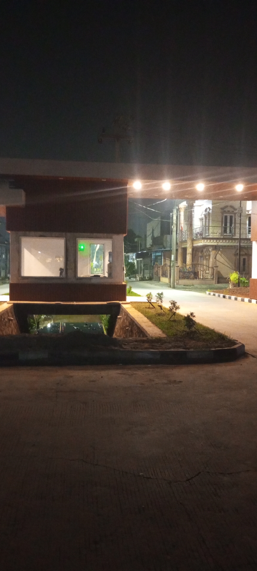 The image depicts a nighttime scene at what appears to be the entrance to a residential complex or security checkpoint. In the foreground, a paved road with visible expansion joints and a slight crack runs across the frame. To the left, a dark, recessed area, possibly a drainage channel or underpass, is visible with some foliage and reflections suggesting water.
The central focus is a small, modern security booth with a flat roof supported by pillars. The booth has two large, rectangular windows. The left window is illuminated from within, showing a blank white surface. The right window is also lit from within and features a bright green light, possibly a status indicator, and a water dispenser. The exterior of the booth has a dark brown textured facade.
Beyond the security booth, a road curves to the right, illuminated by overhead lights. On the right side of the road, a multi-story building with an ornate facade, balconies, and numerous windows is visible. This building exhibits a classical architectural style. In the middle ground, between the security booth and the building, there are fences, smaller structures, and some planted greenery with small trees. The overall environment suggests an urban or suburban setting at night. The scene is captured from a low angle, looking slightly upwards towards the security booth and the structures beyond.