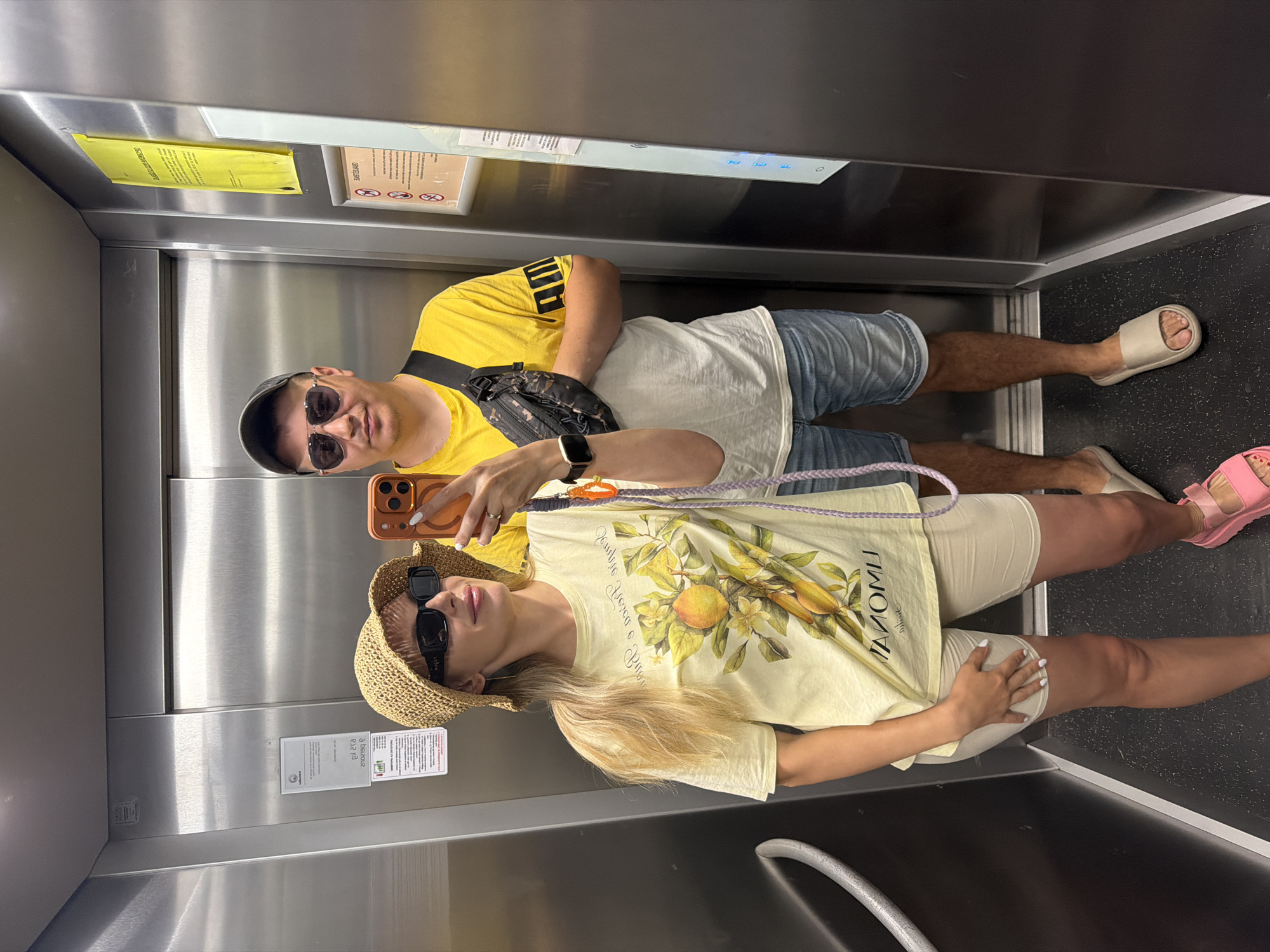 Inside an elevator, two people, likely a couple, are taking a selfie. The man is wearing a yellow shirt, blue shorts, and a black fanny pack. The woman is wearing a light yellow t-shirt, light shorts, a straw hat, and pink sandals. The elevator is lined with metal, and there are informational signs on the wall. The location is Budva, Montenegro.