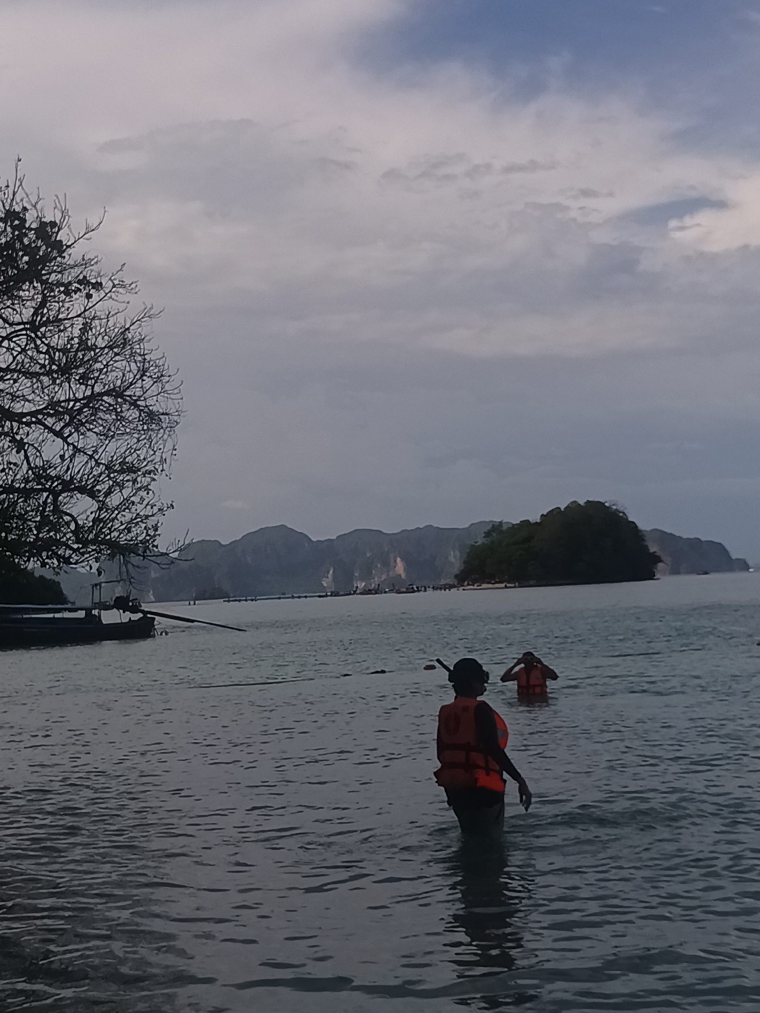 The image depicts a coastal scene in Ao Nang, Thailand. The water is murky and grey. Two people wearing orange life jackets are wading in the water. The person closest to the viewer is facing away, wearing a snorkeling mask. In the background are islands, one covered in trees, with low-lying mountains further behind. To the left, a bare tree and the side of a boat are visible. The sky is overcast with grey clouds.