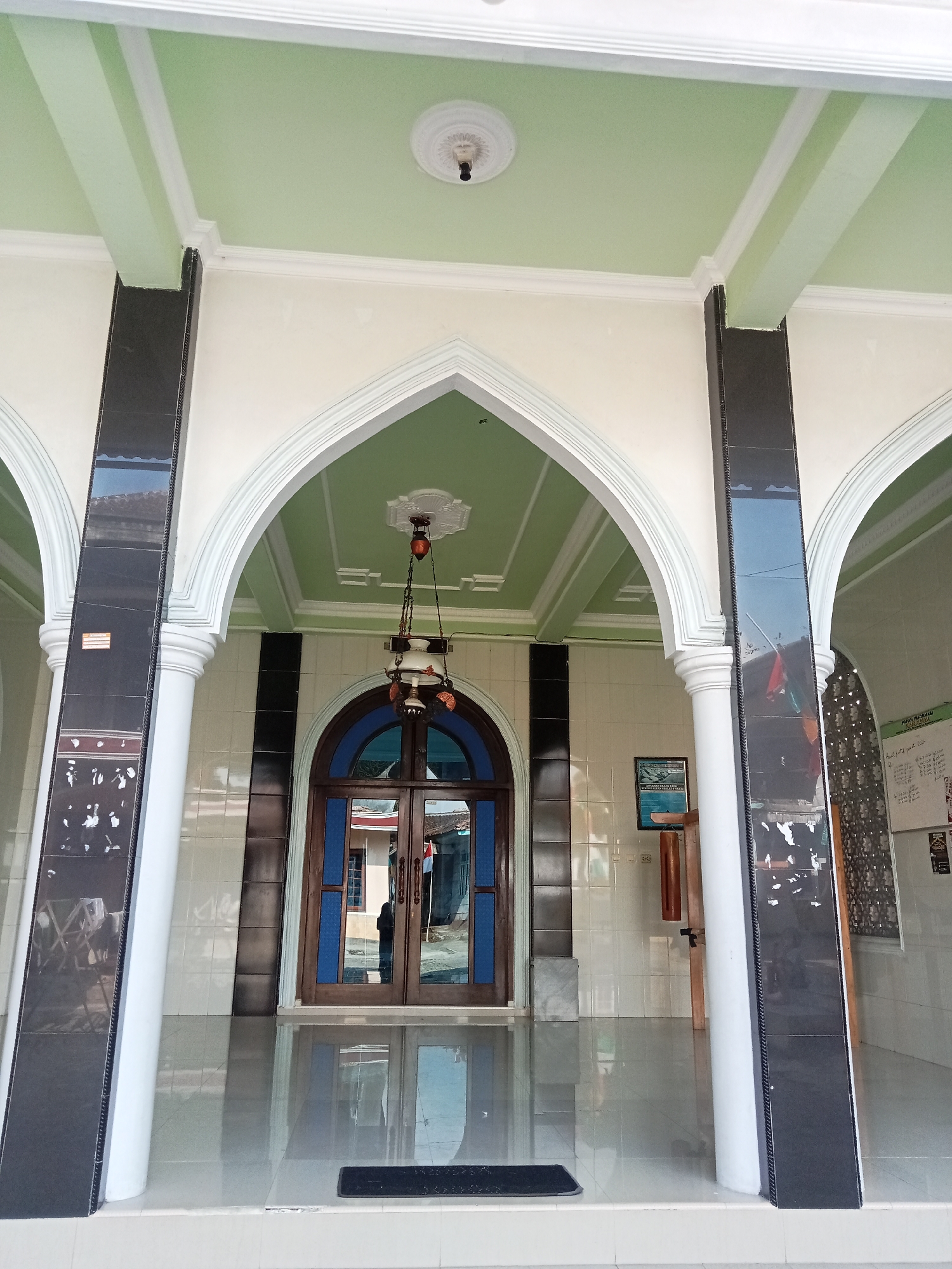 The image shows the entrance to a building, likely a mosque, in the city of Pancurwening, Indonesia. The entrance features a large arched doorway with double wooden doors. Above the doors, a decorative chandelier hangs from the ceiling. The exterior of the building is characterized by white pillars, dark decorative tiles, and a light green ceiling. The floor is tiled and reflects the architectural elements. There is a person visible through the glass doors, and other architectural details like posters on the wall are present. The overall scene depicts a clean and well-maintained building entrance.
