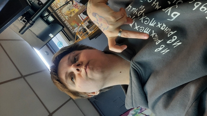 An individual with short brown hair, wearing a dark gray hooded sweatshirt featuring white Cyrillic text, is depicted indoors at a low angle in Saratov, Russia. The person has a septum piercing, two eyebrow piercings on the left brow, and multiple ear piercings on the left earlobe. A small, faint heart-shaped mark is present on the upper left cheekbone, and a mole on the lower neck. The subject's lips are pursed, and their gaze is directed upwards and slightly to the right. The right hand is raised, with the index and pinky fingers extended while the middle and ring fingers are bent, forming a gesture. A silver-colored ring is worn on the right ring finger, and a faint, light-colored tattoo resembling a cartoon character is visible on the back of the right hand. The background features a white suspended ceiling with a grid pattern, overhead fluorescent lighting, and a recessed circular light. A black Philips computer monitor is visible on the upper right, mounted on an arm, with cables connected to its rear. Below the monitor, a white air conditioning unit is present. A partially obscured sign in Russian, "УГОЛОК ПОТРЕБИТЕЛЯ" (Consumer's Corner), is visible, confirming a retail or service establishment. A wooden display rack and abstract wall graphics are also discernable in the background.
