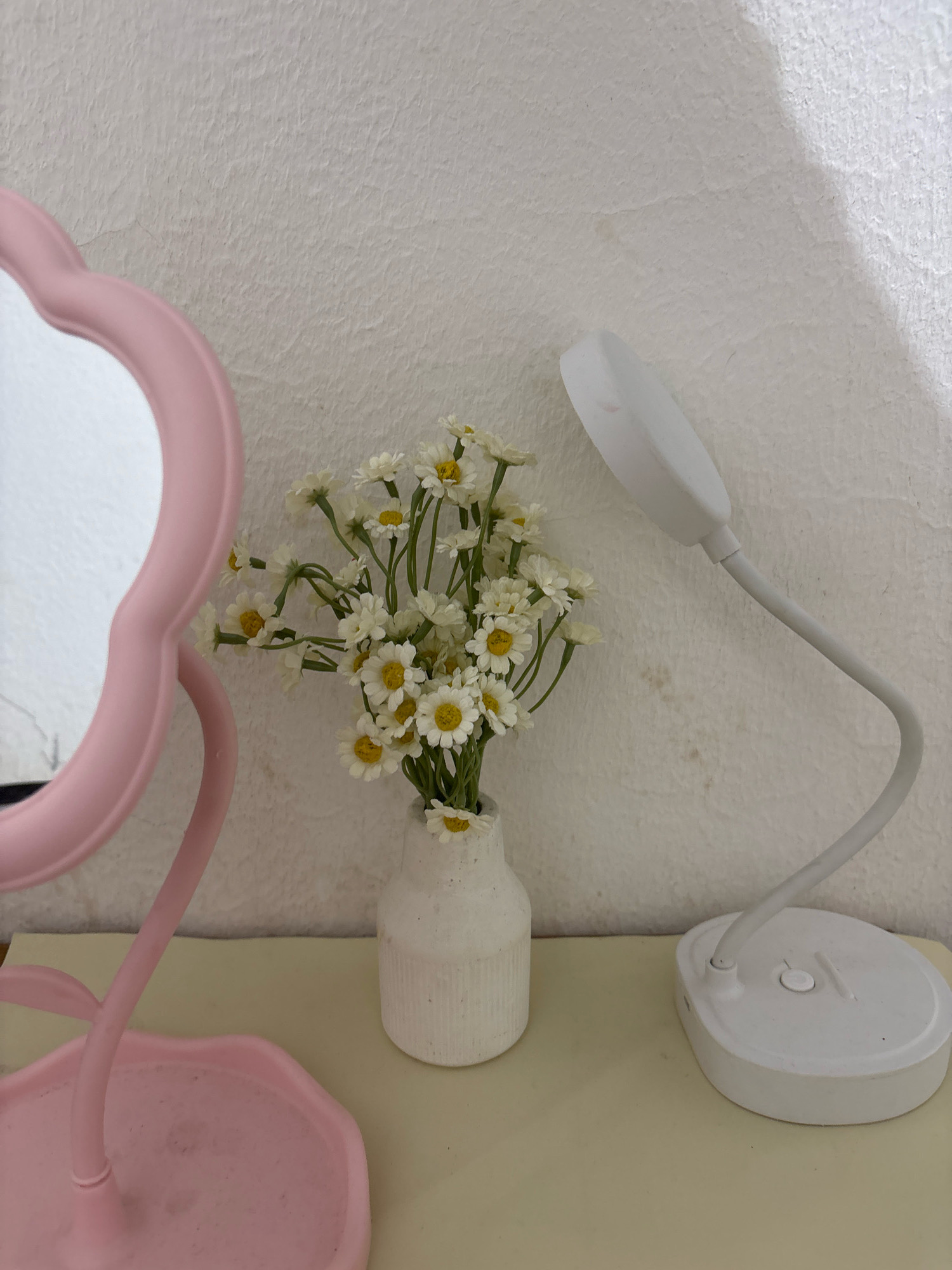 The image depicts a still life arrangement on a pale yellow surface, likely a tabletop or shelf, against a textured white wall. The scene features a pink decorative mirror with a scalloped edge on the left, a small white vase filled with an arrangement of white daisy-like flowers with yellow centers in the middle, and a white desk lamp with a flexible gooseneck on the right. The mirror has a delicate stand and base. The flowers are clustered and appear to be artificial. The desk lamp has a circular base with a single button on its surface. The overall impression is one of simple decoration or a personal vanity area. The location in the city of Wanajaya, Indonesia is not evident from the image itself.