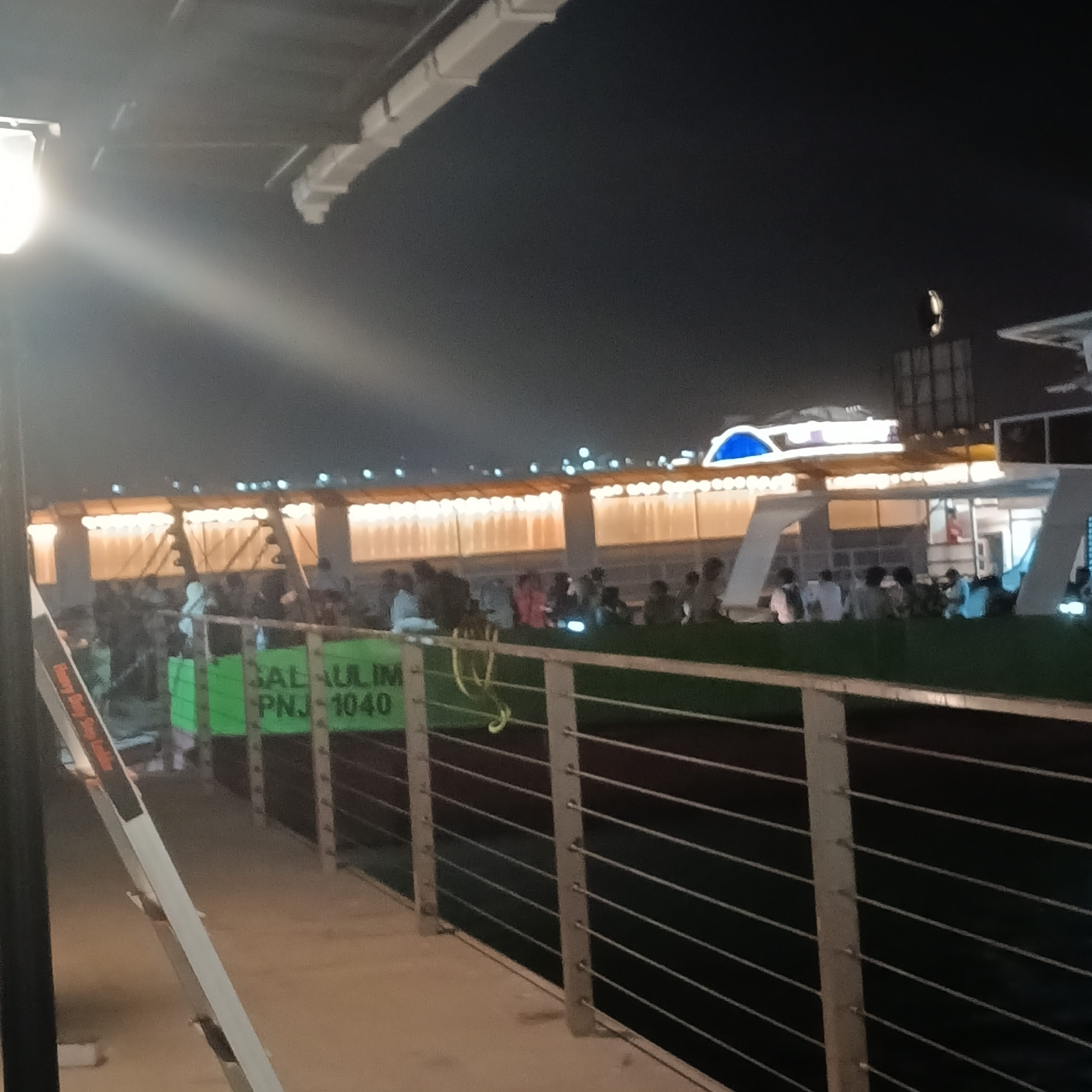 The scene is set at night in Panaji, India, with a view overlooking what appears to be a body of water. A large crowd of people is gathered at a building in the distance, illuminated with numerous lights. The building has a blue dome-like structure on top. In the foreground, there is a railing made of horizontal bars and vertical posts. To the left, there's a light post casting a bright light. A green sign with "SAL GAULIM-PNJ-1040" is visible behind the railing, partially obstructed by other people. The overall environment is dark, with artificial lighting defining the scene.