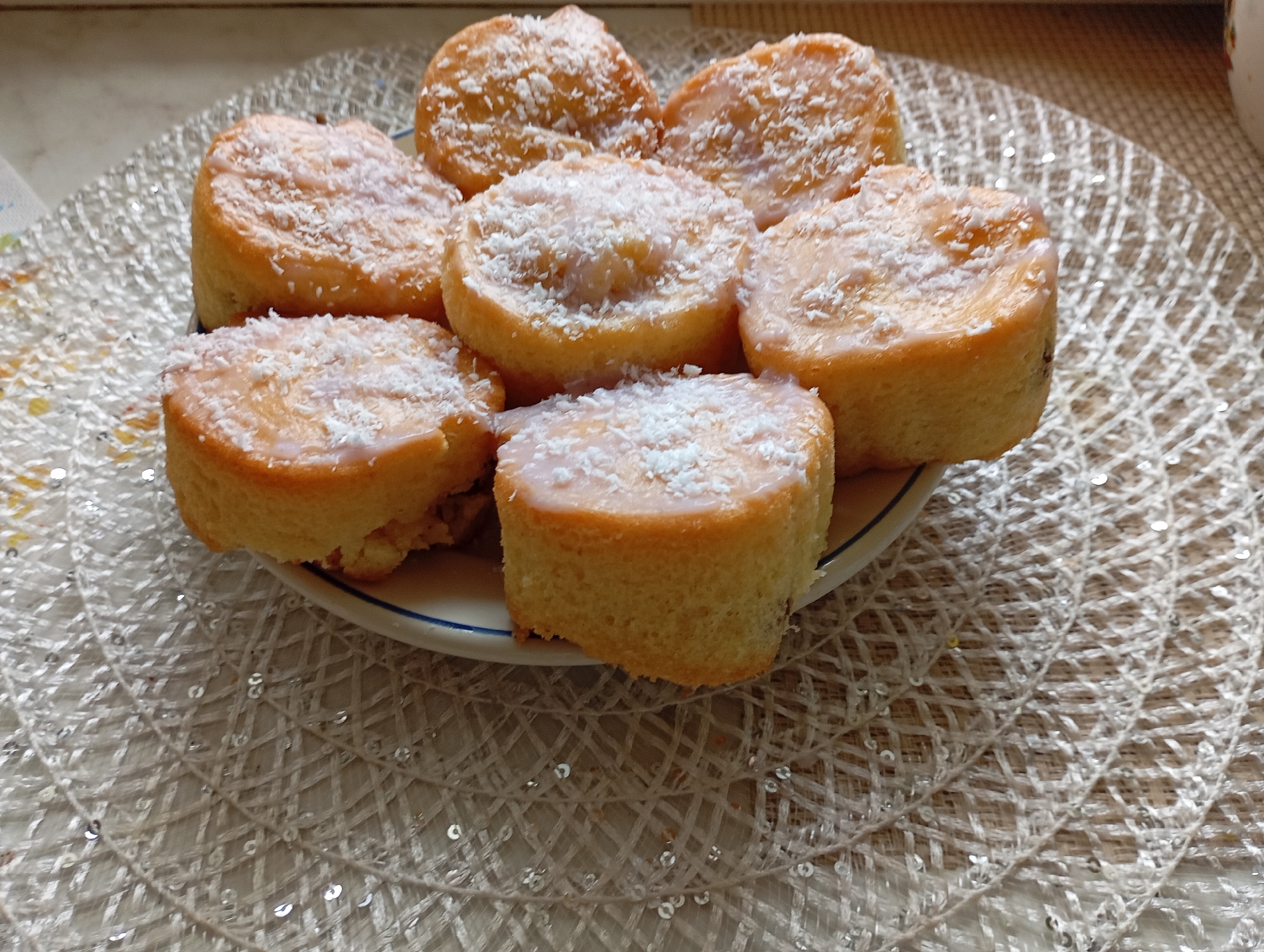 The image shows a plate of seven small, round cakes topped with a light glaze and sprinkled with shredded coconut. The cakes are arranged closely together on a plate with a blue rim. The plate is placed on a round, silvery placemat with a woven texture. The scene is brightly lit, possibly by natural light coming from a window. The location is likely indoors. Given the request to include the city of Bydgoszcz, Poland, it can be assumed that this image was taken in a household or bakery in Bydgoszcz.