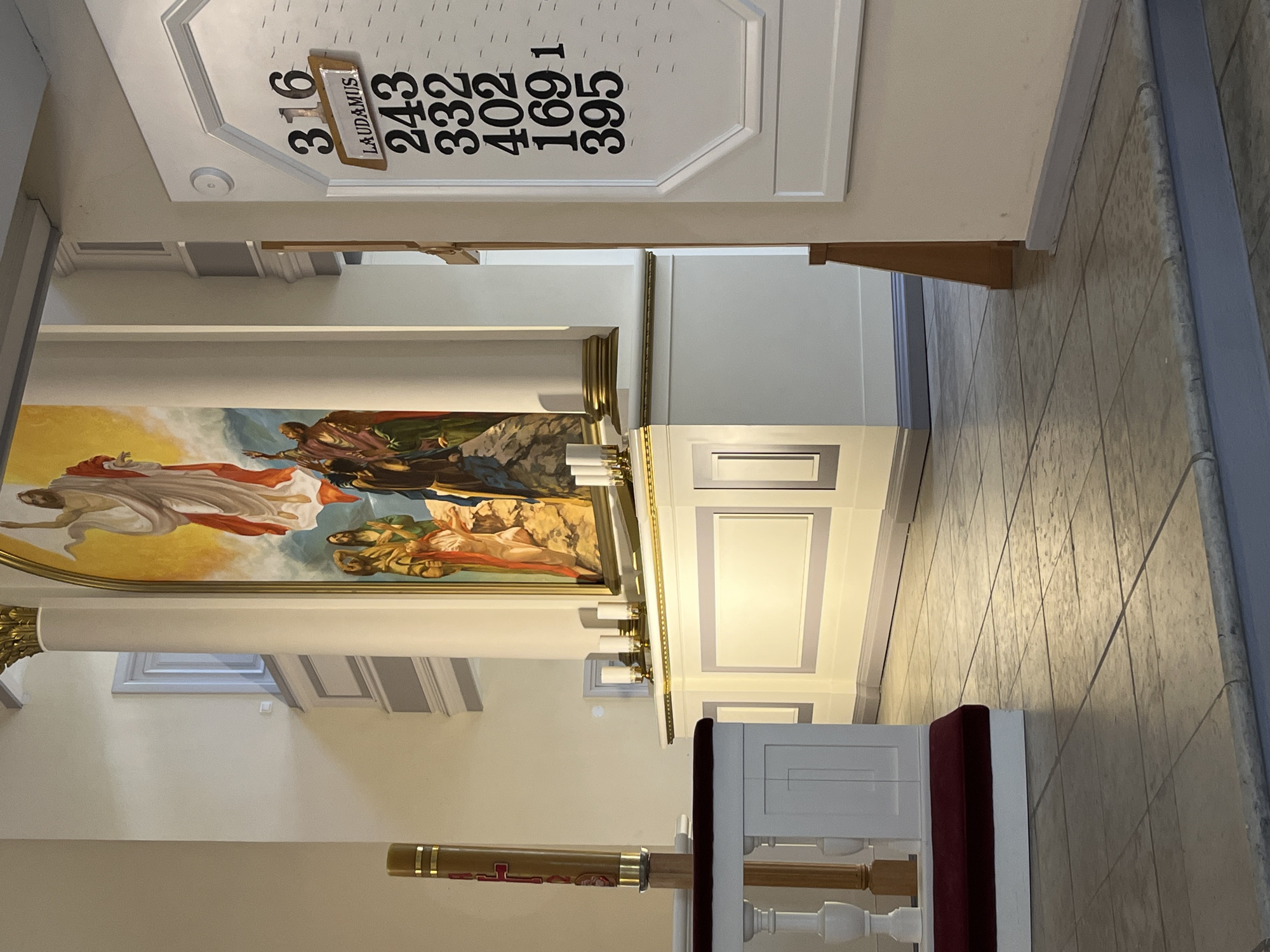 This is an interior shot of a church in Saint Petersburg, Russia. Visible are the ceiling displaying hymn numbers "316, 243, 332, 402, 169, 395", a large mural depicting a biblical scene, a pulpit with a red cushion, and architectural details such as columns and tiled flooring. The environment appears to be well-lit and maintained. There are no people visible in the image.