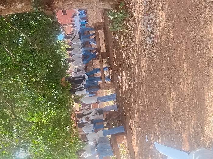A line of seated individuals in white shirts and blue pants or skirts, possibly students, are visible in the image. They are sitting on a wooden structure in an outdoor setting. The area is under a tree, and the ground appears to be dirt or sand with some debris scattered around. A building is visible in the background. The photo is taken from a slightly elevated angle and the orientation is rotated.